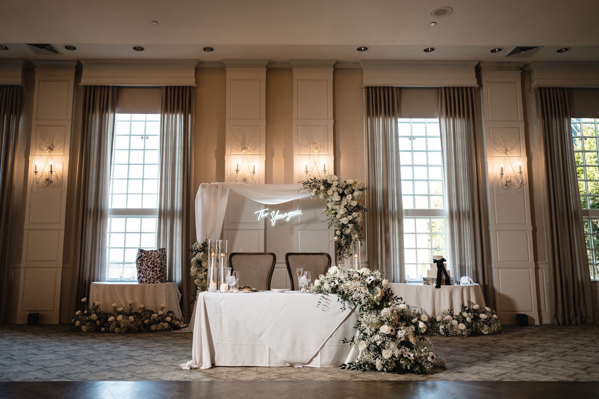 Elegant wedding sweetheart table adorned with white floral arrangements and a draped backdrop in a ballroom.