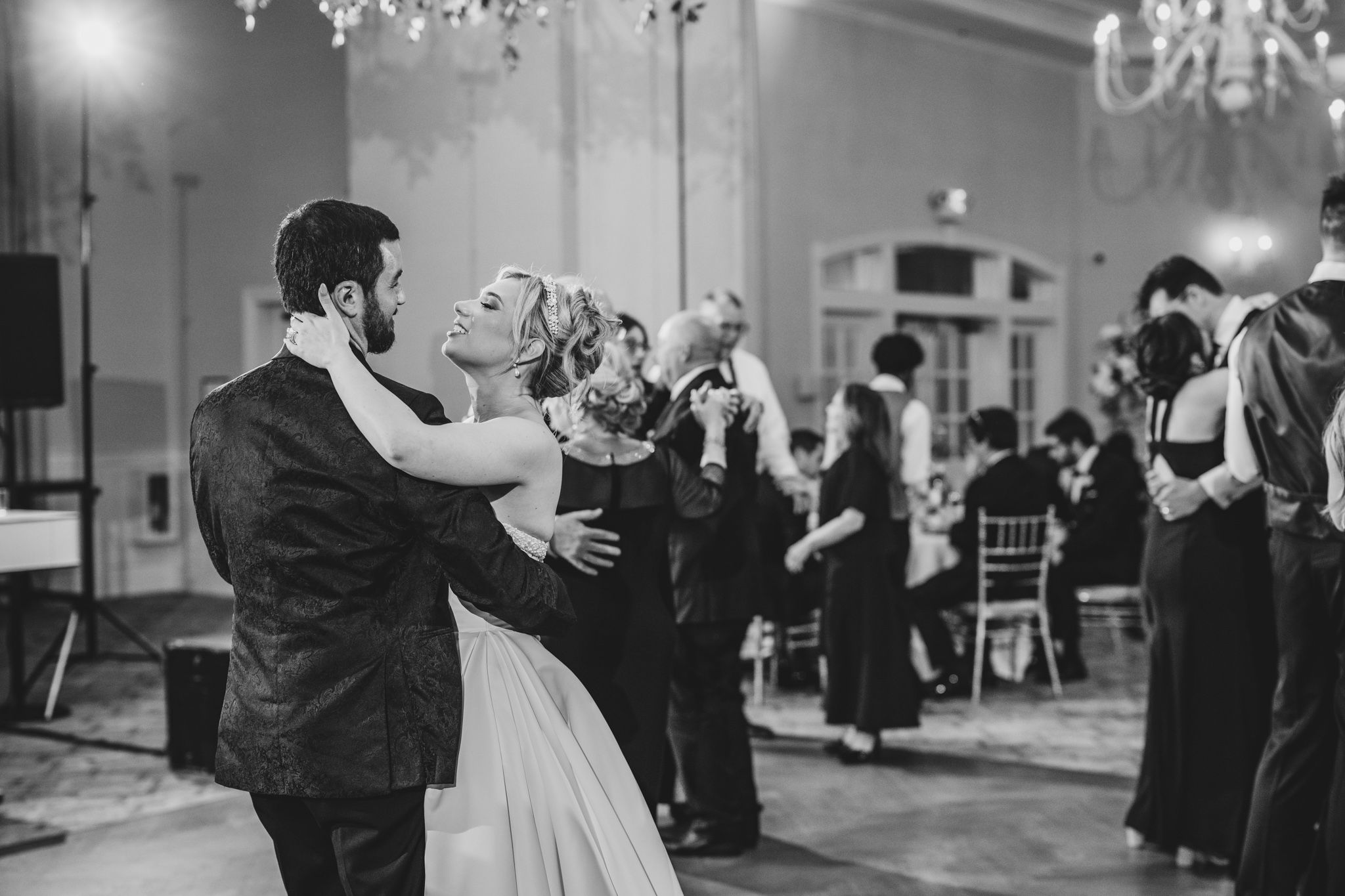 Black and white photo of bride and groom sharing an intimate slow dance at their wedding reception with guests dancing.