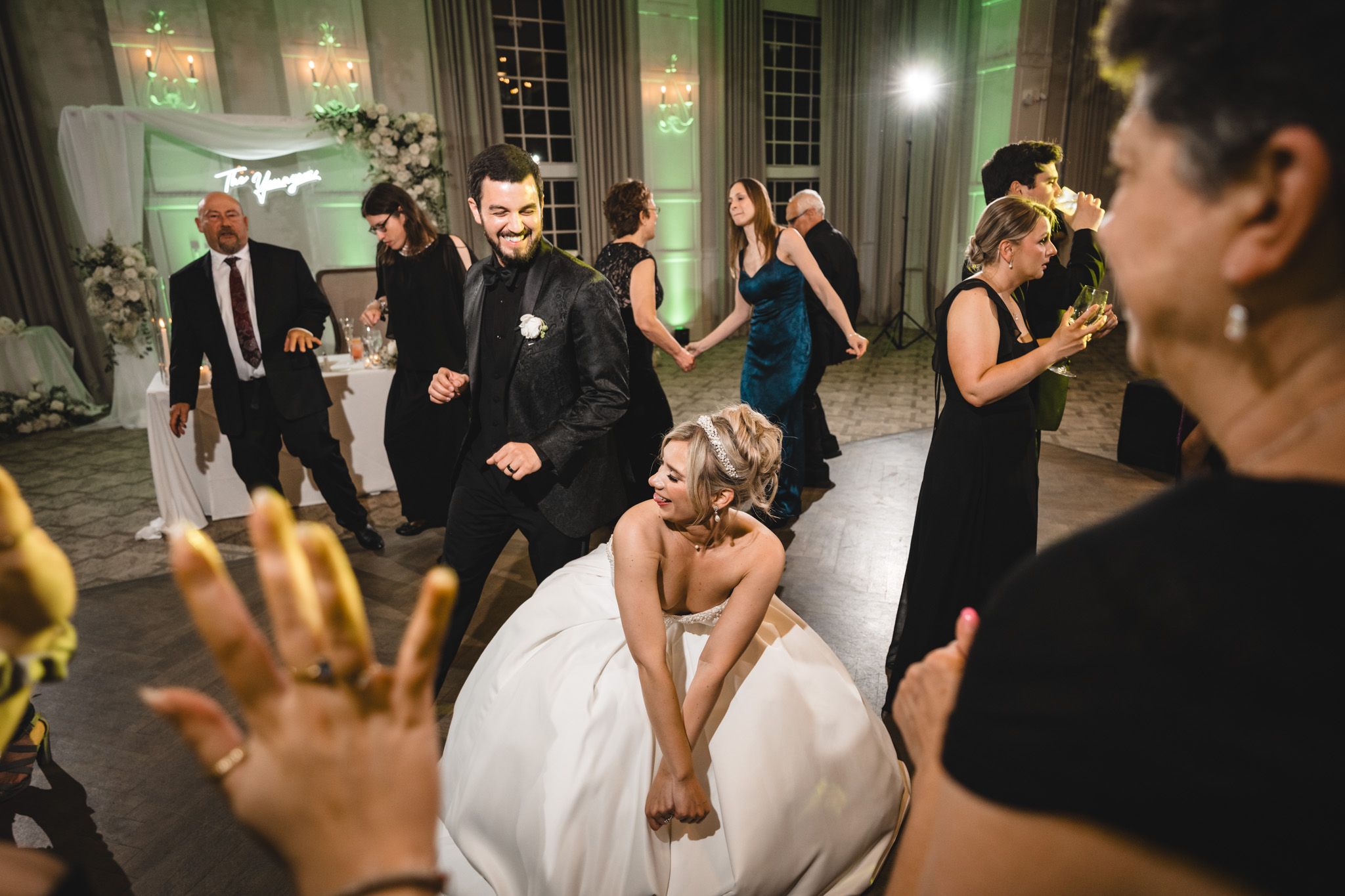 Bride in white gown sits on dance floor laughing during lively wedding reception celebration.