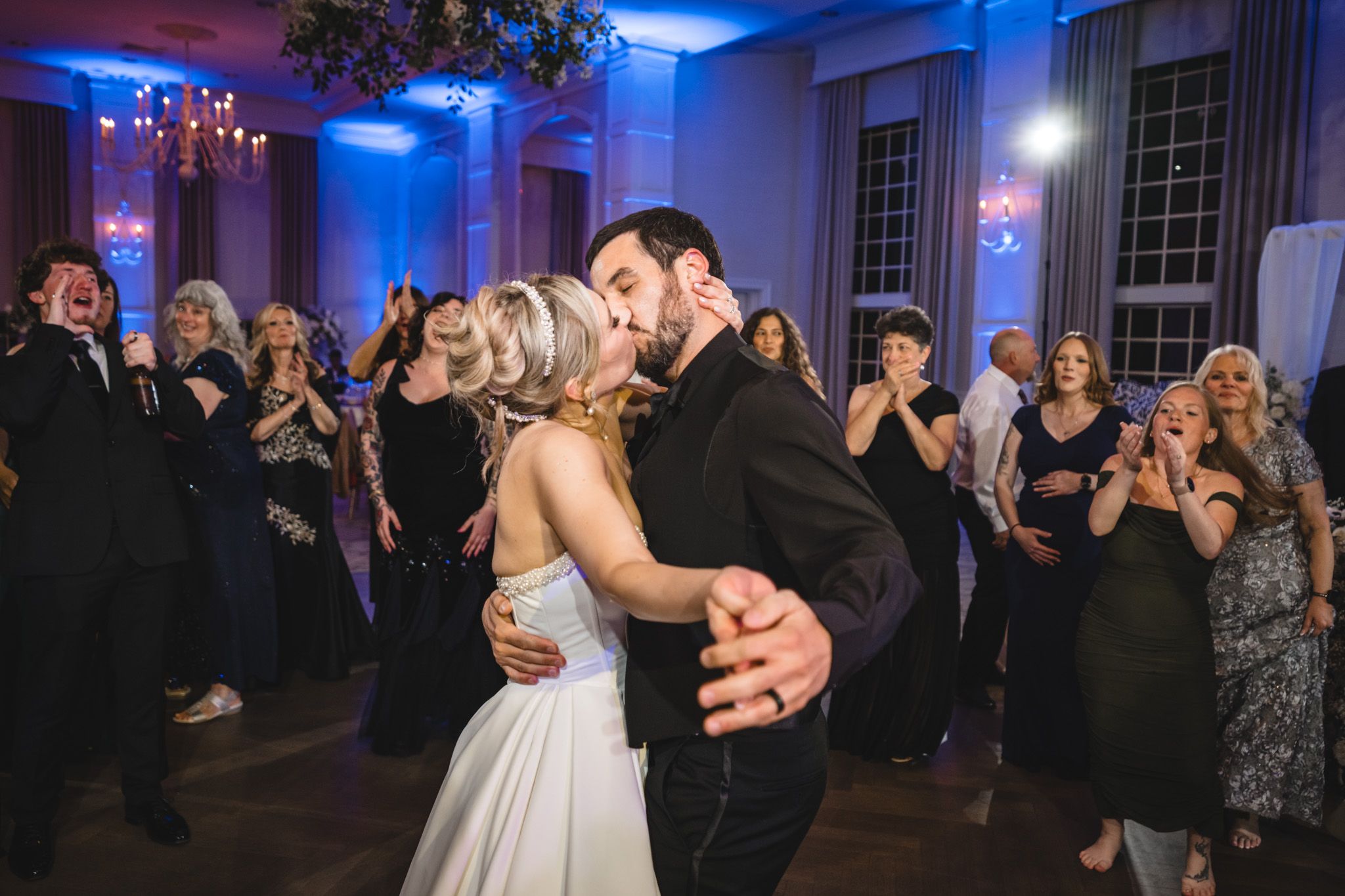 Bride and groom share romantic first dance kiss surrounded by cheering guests at elegant wedding reception.