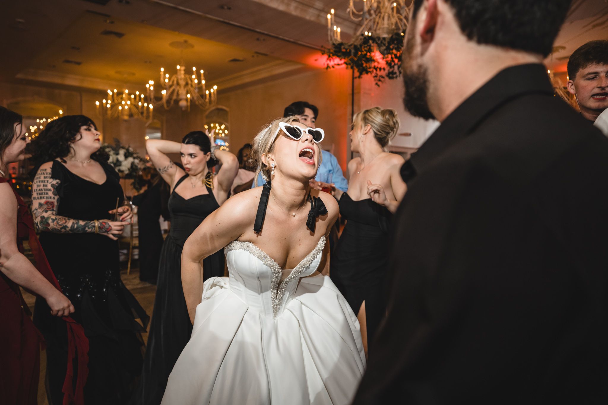 Bride wearing white sunglasses dances energetically with guests at lively wedding reception celebration.
