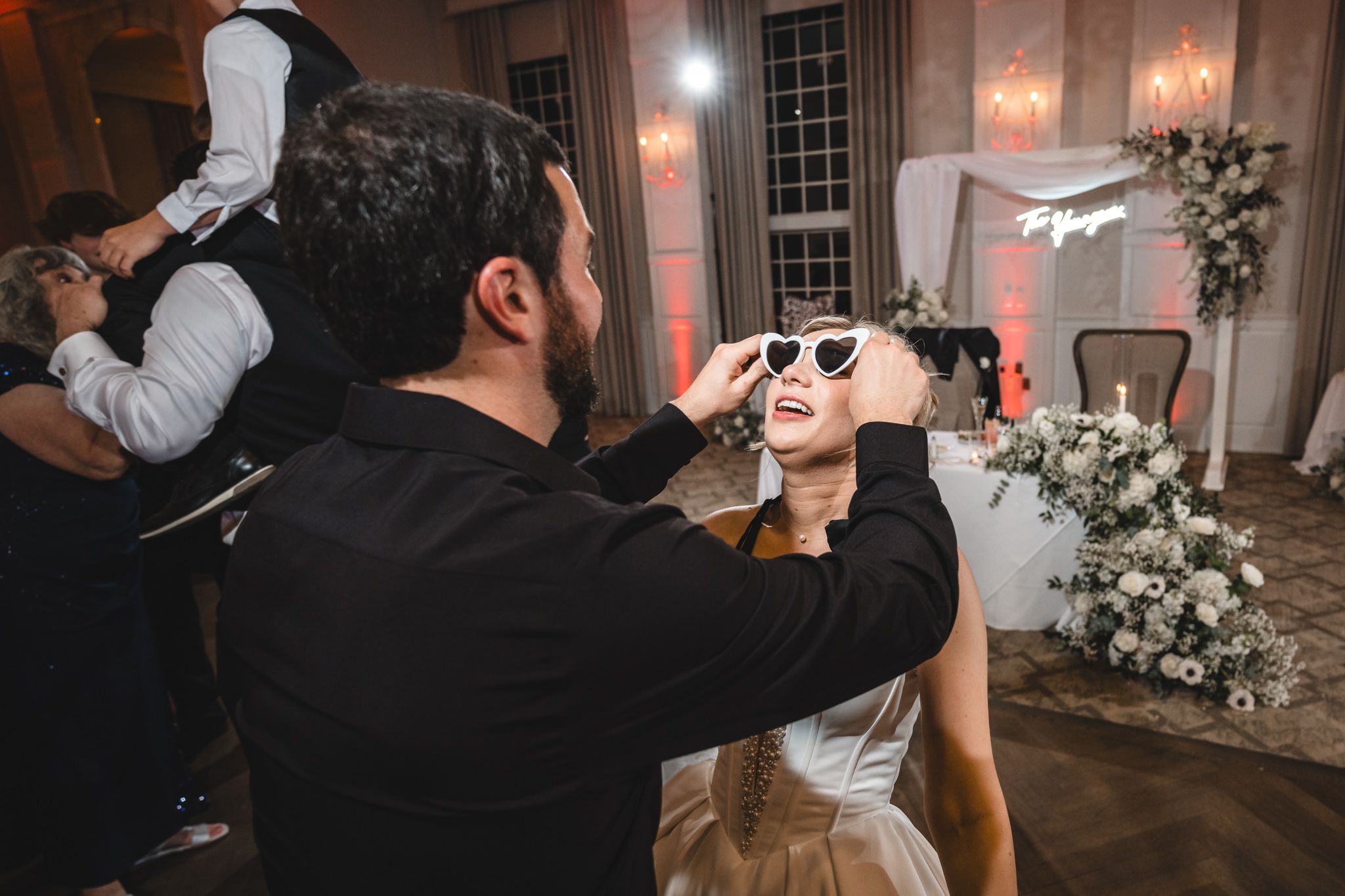 Groom places heart-shaped sunglasses on laughing bride during fun wedding reception celebration.