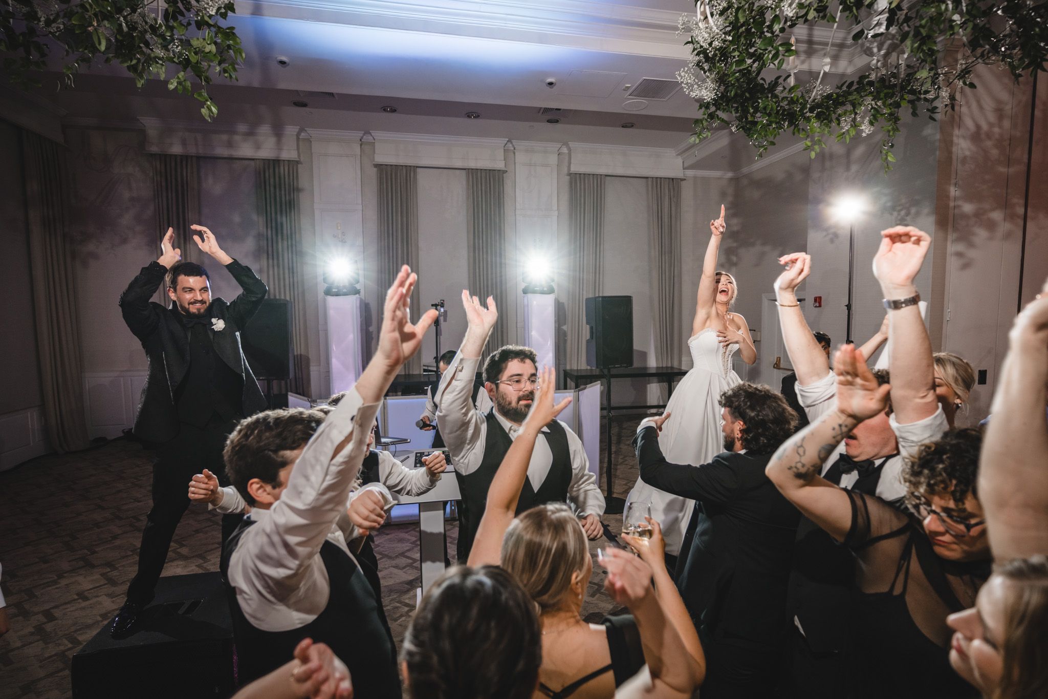 Bride, groom, and wedding guests celebrate together with hands raised on the reception dance floor.