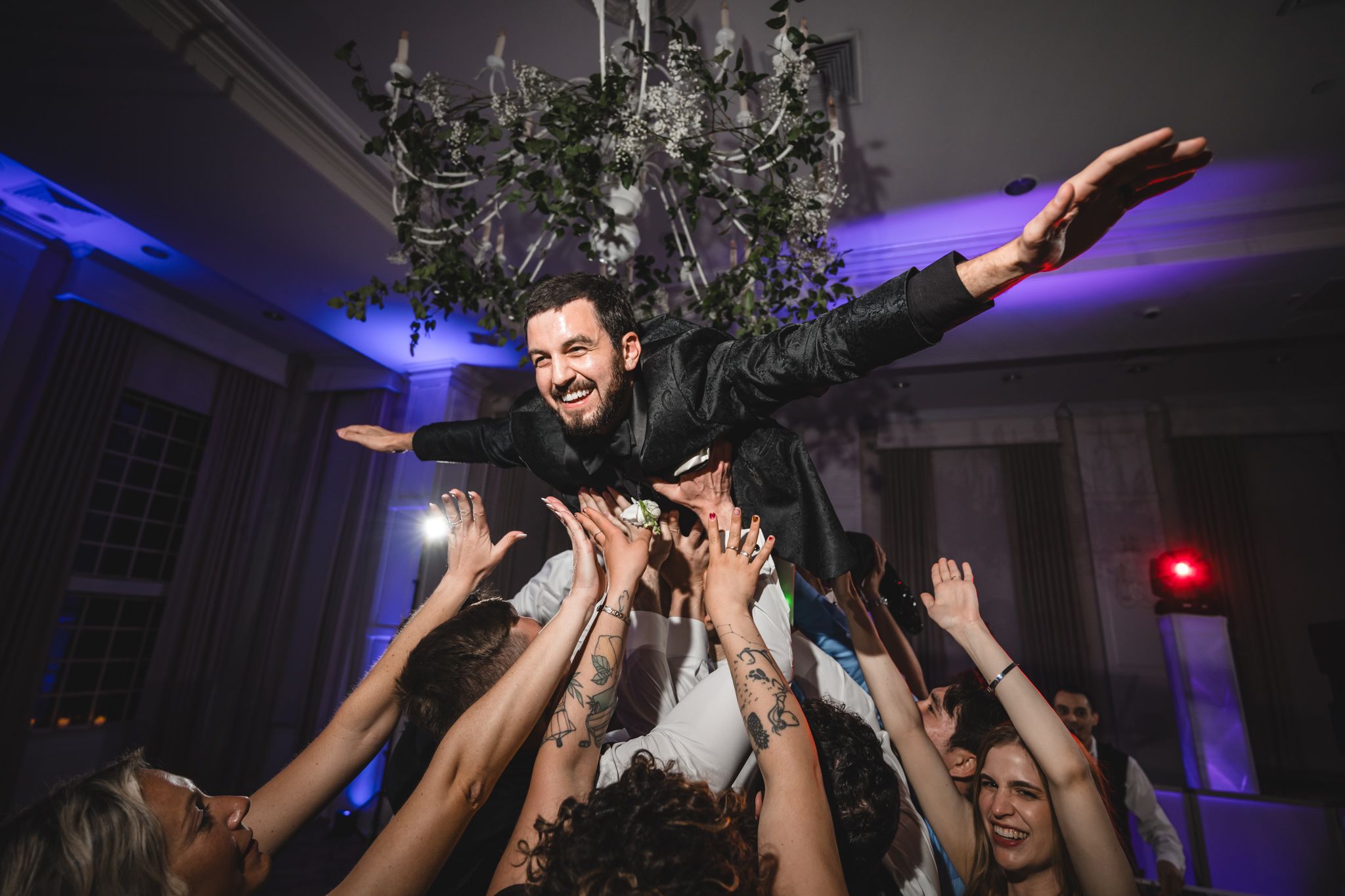 Groom being lifted by wedding guests in a celebratory crowd surf moment during lively indoor wedding reception.