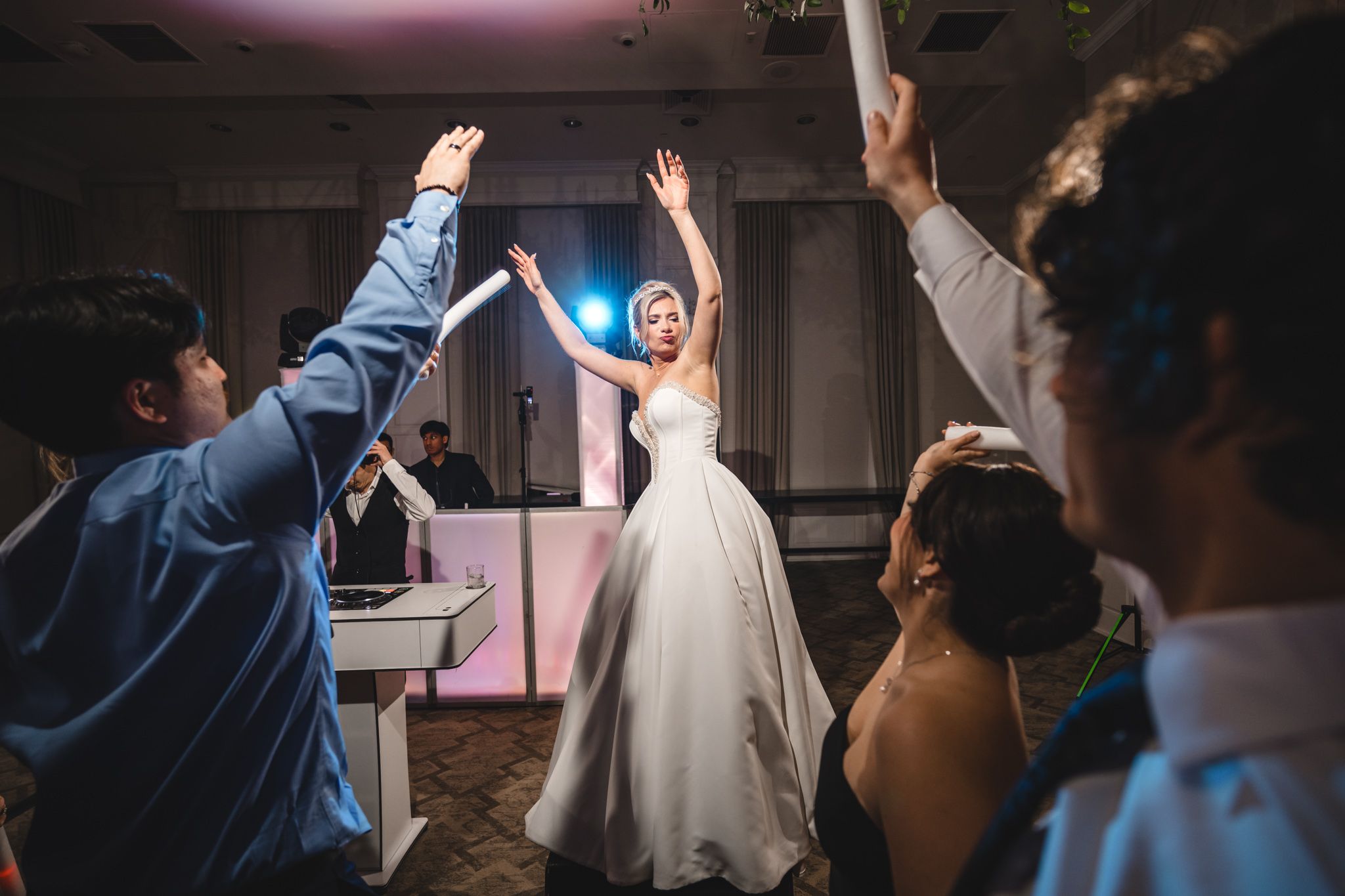 Bride in strapless white gown dancing joyfully with raised arms surrounded by cheering guests at wedding reception.