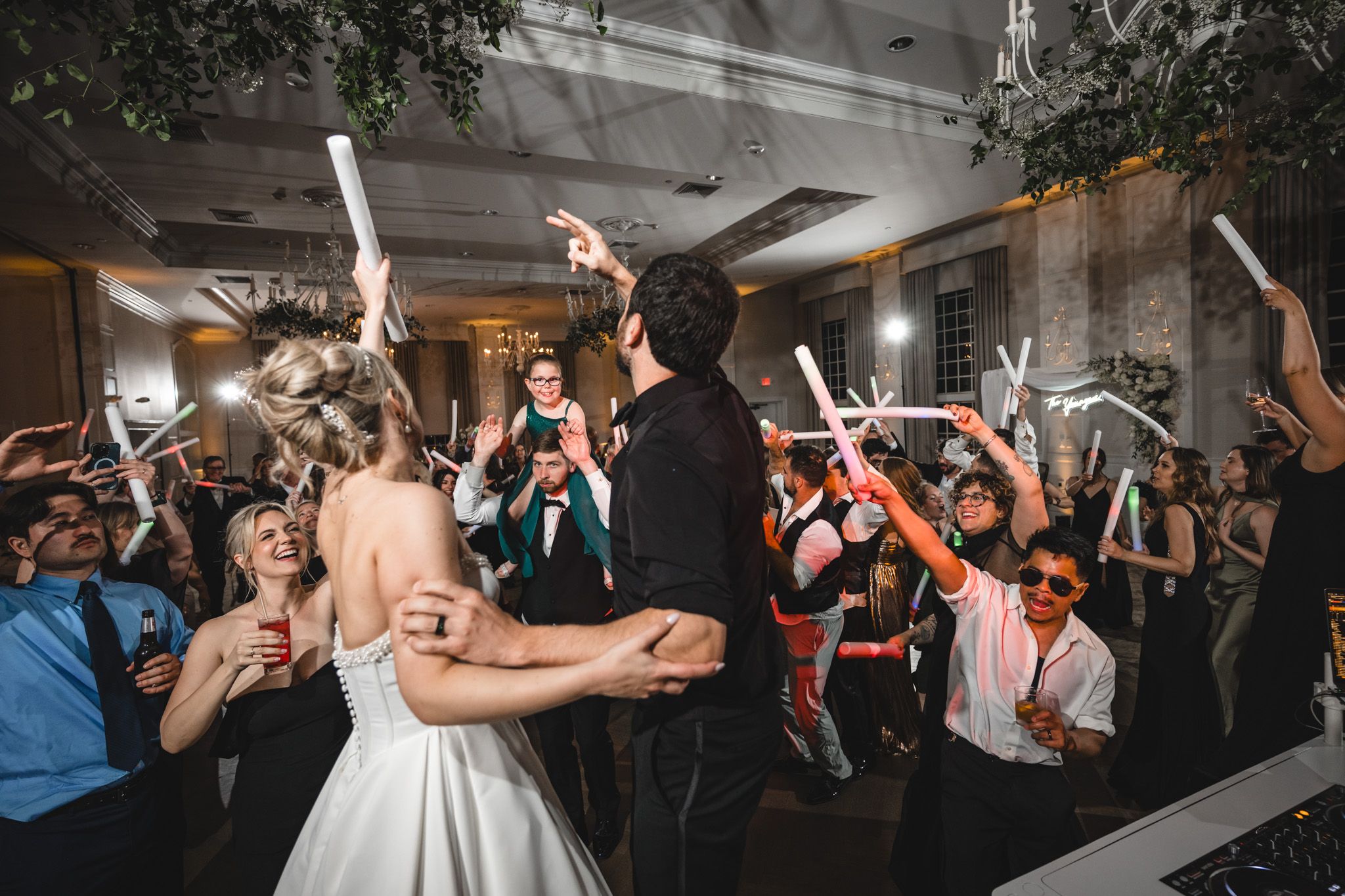 Bride and groom dance surrounded by cheering guests waving glow sticks at lively wedding reception.