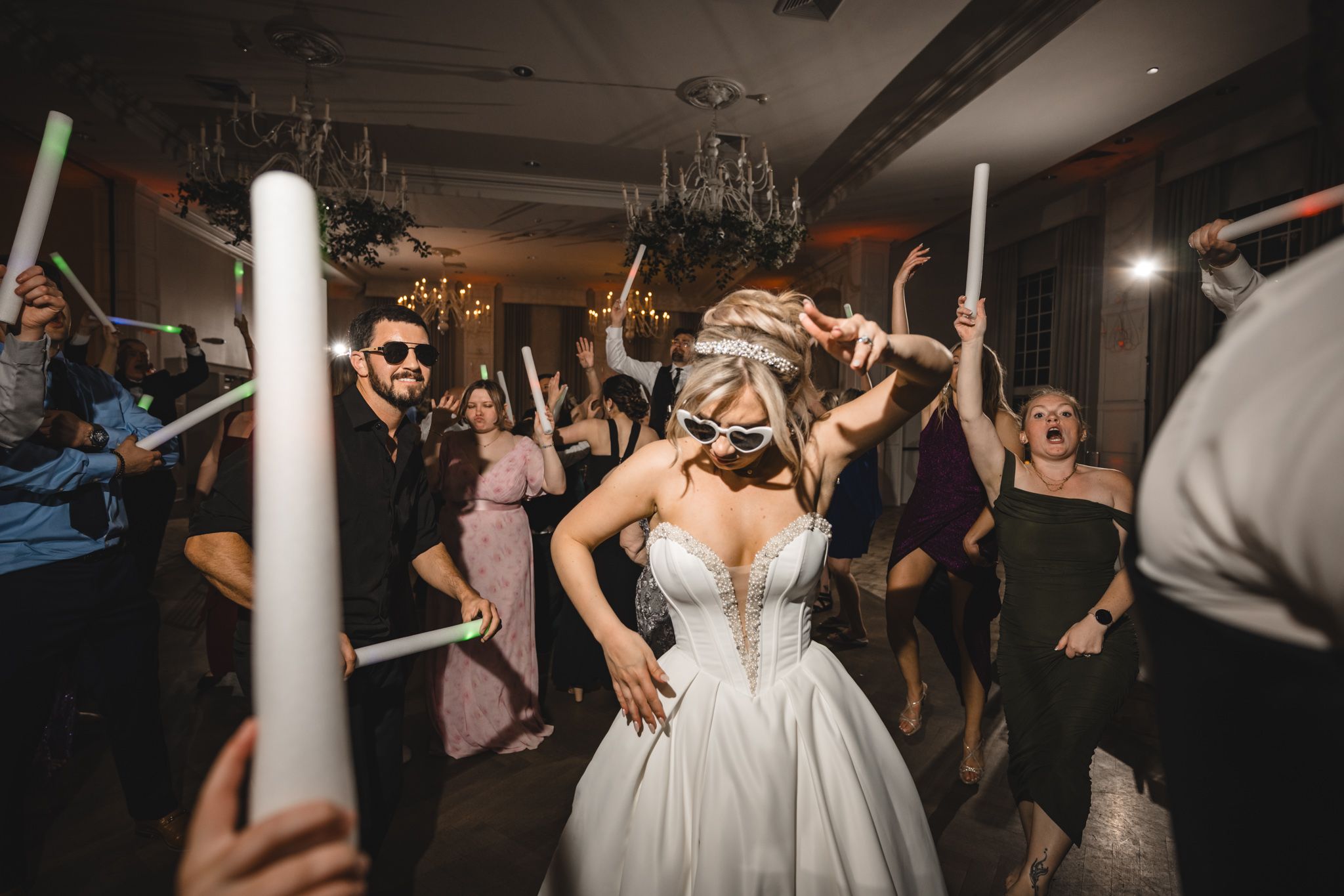 Bride in white gown dances joyfully with guests holding glow sticks at lively wedding reception party.