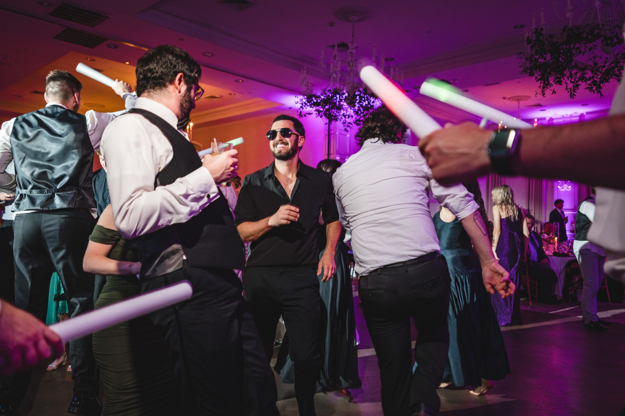 Guests dancing energetically with glow sticks under purple lights at a festive and lively wedding reception party.