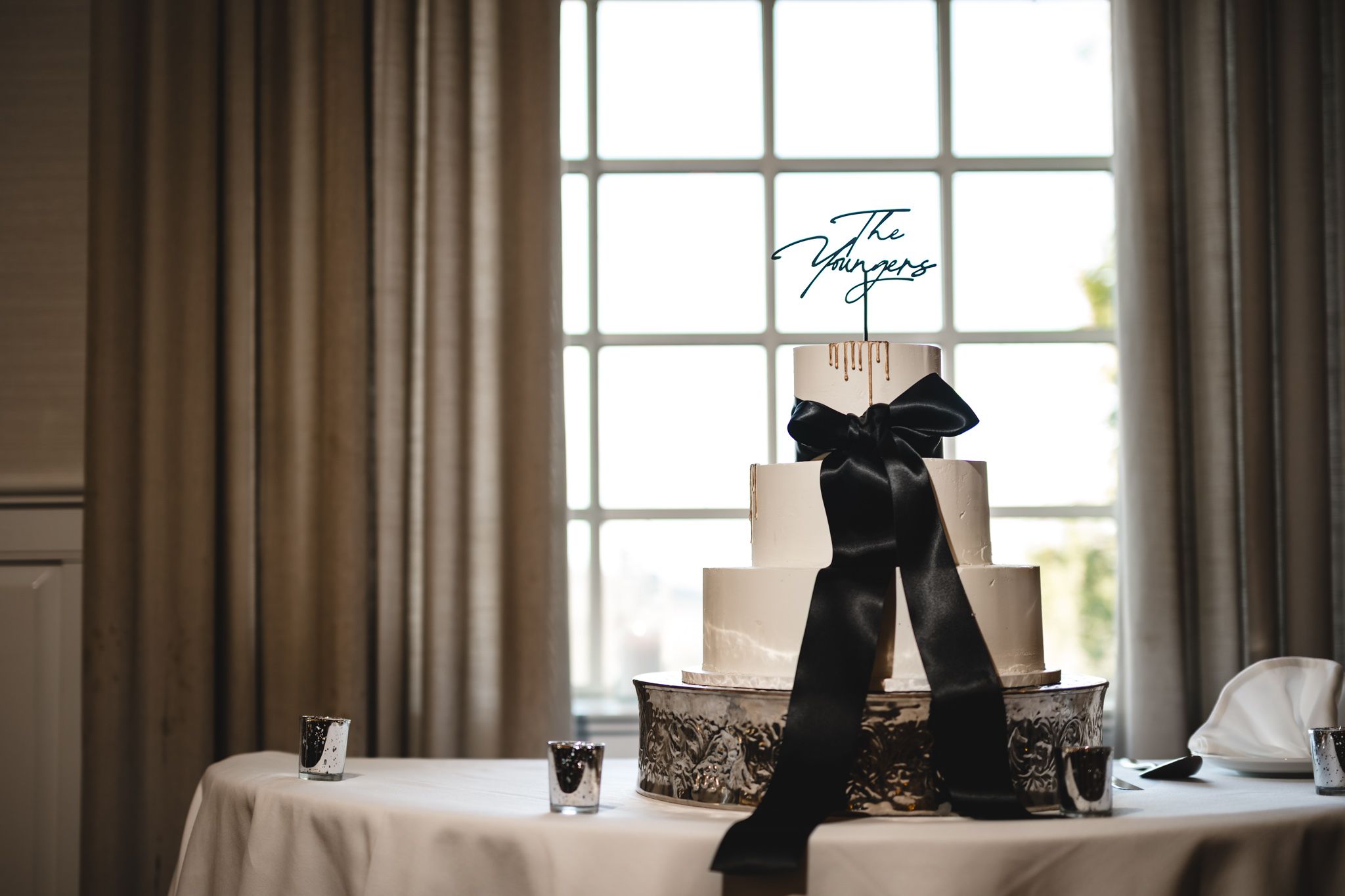Elegant white wedding cake with black satin bow and personalized topper displayed on a decorated table.