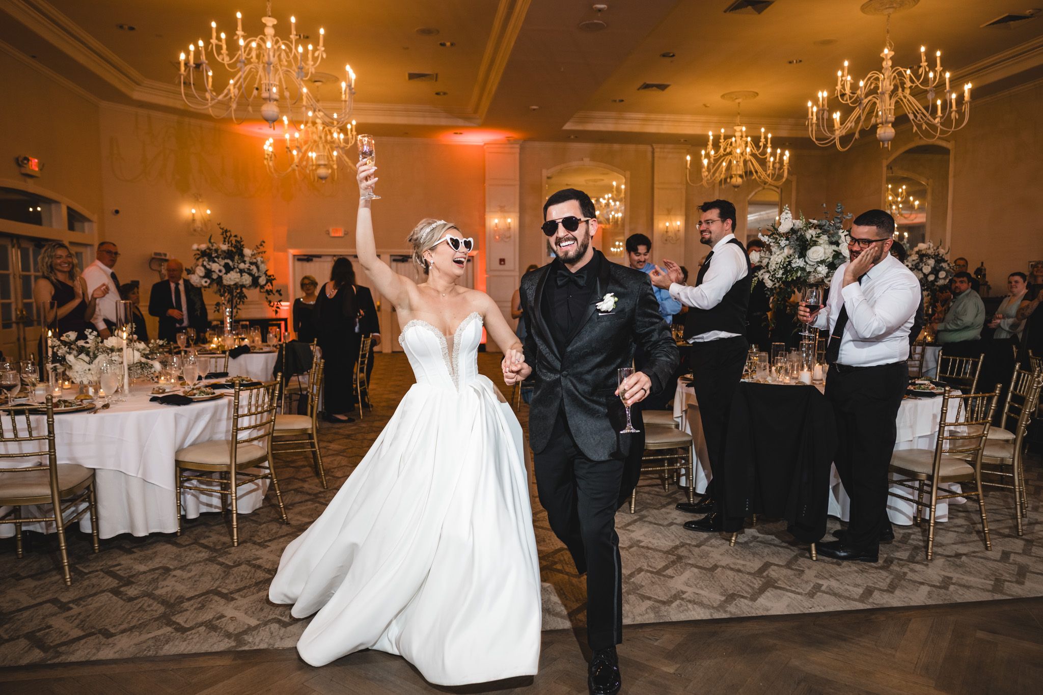 Bride and groom make grand entrance into elegant wedding reception hall with chandeliers and floral centerpieces.