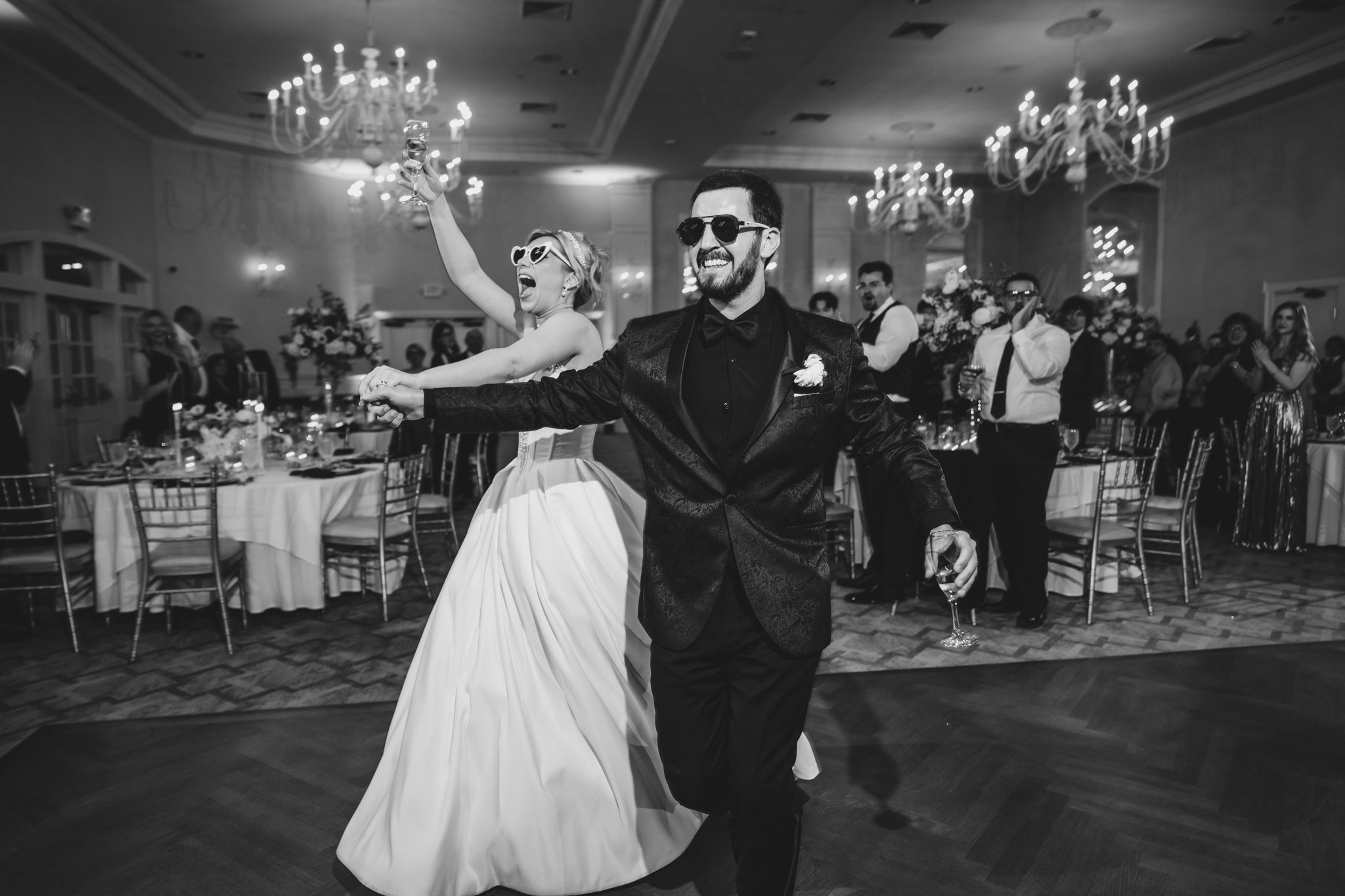 Black and white photo of bride and groom sharing a fun first dance, groom wearing sunglasses in elegant ballroom.