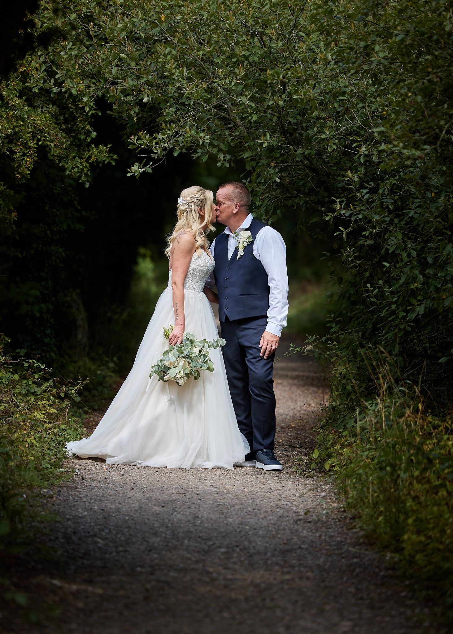 Bride and groom sharing a romantic kiss on a tree-lined path with soft natural lighting.