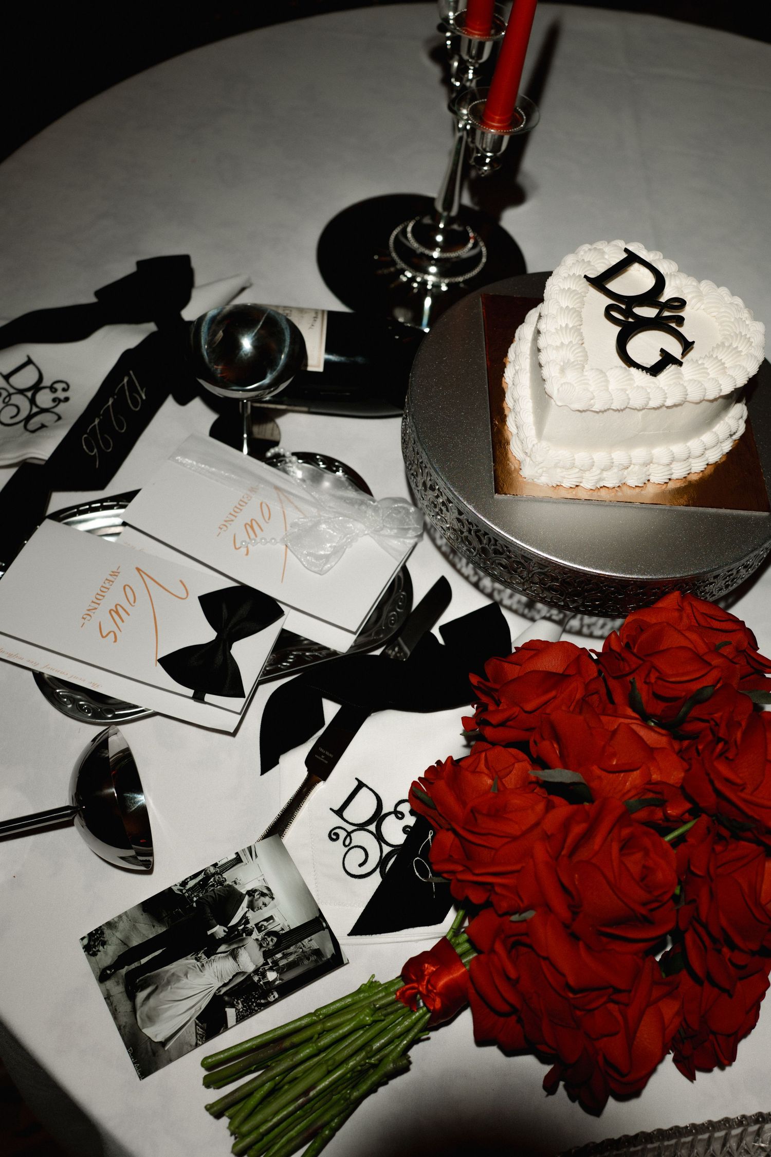 Elegant wedding table with heart-shaped D&G cake, red roses bouquet, black bow cards, and silver candlesticks on white tablecloth.