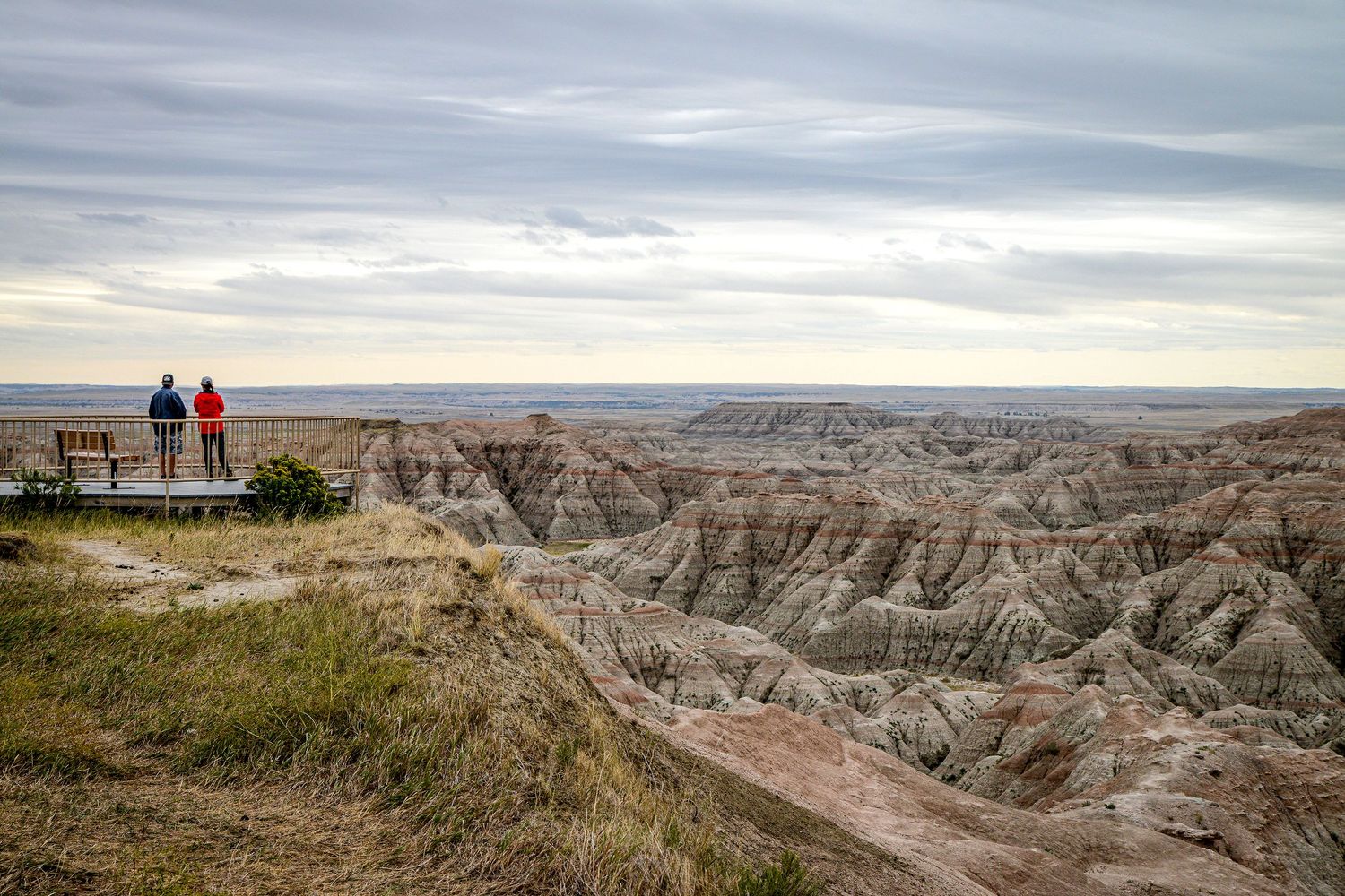 Two visitors stand at a scenic overlook admiring the vast eroded rock formations of Badlands National Park under cloudy skies.