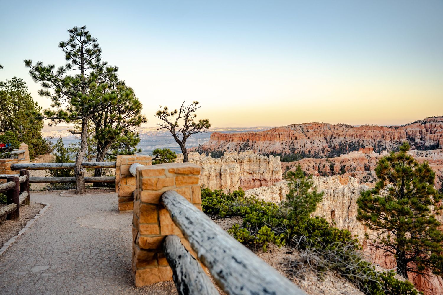 Scenic Bryce Canyon overlook at sunset with wooden railings, hoodoo rock formations, and warm golden light on the horizon.