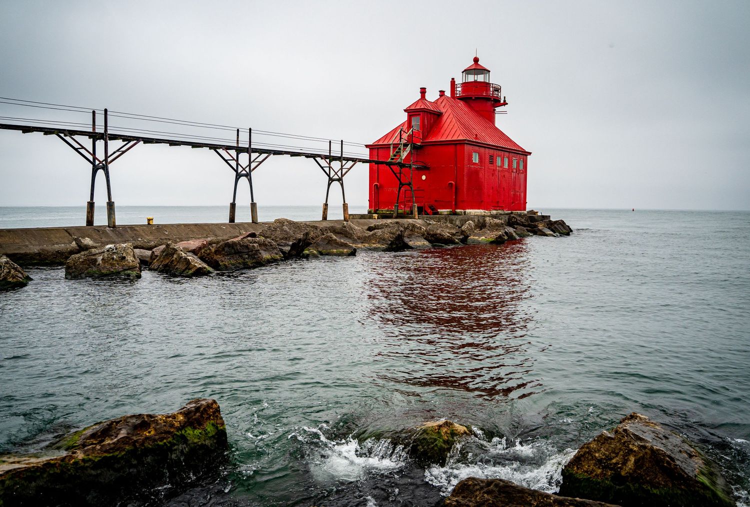 Striking red lighthouse standing on rocky Lake Michigan pier under a cloudy grey sky.