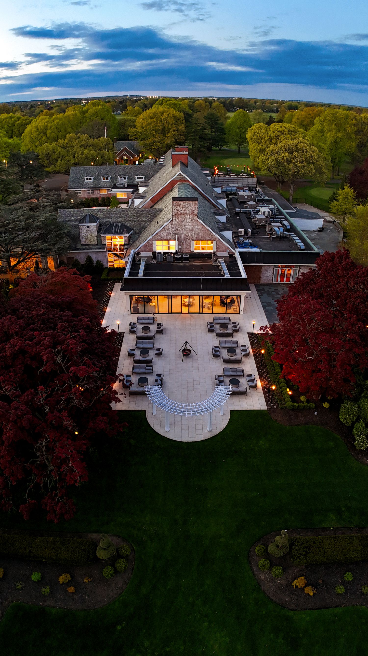 Aerial view of an elegant outdoor patio with white pergola, lounge seating, and a fire pit beside a warmly lit glass building.