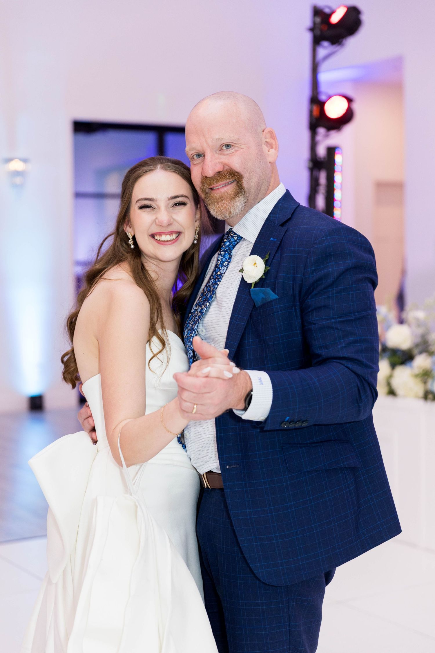 Bride and father share a first dance at wedding reception, both smiling, she in white gown, he in navy suit with boutonniere.