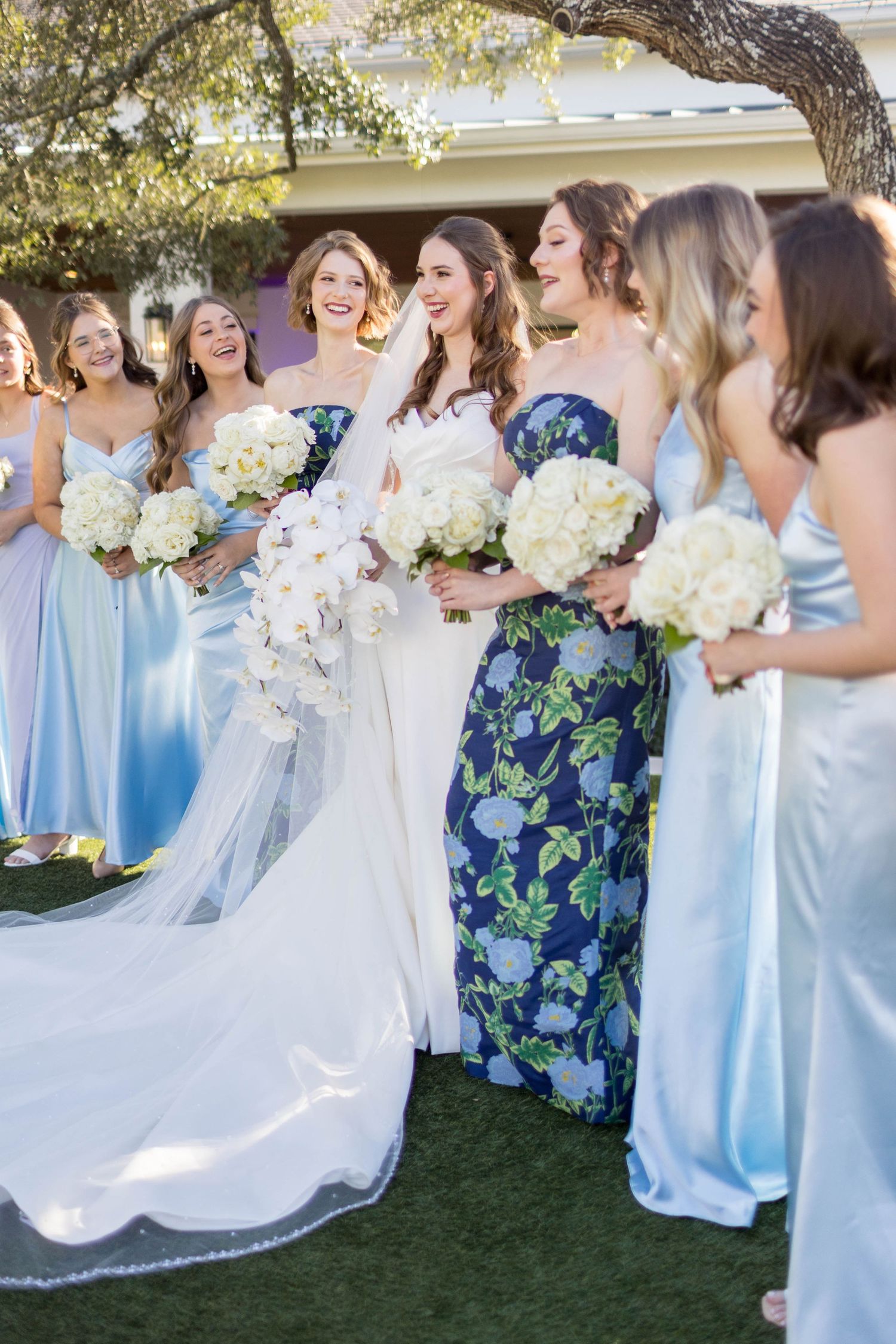 Bride in white gown with bridesmaids in light blue satin and floral dresses holding white hydrangea bouquets outdoors.