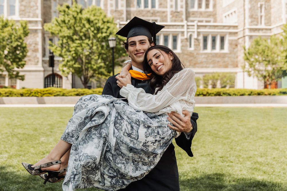 Graduate in cap and gown carrying woman in his arms on sunny college campus lawn.