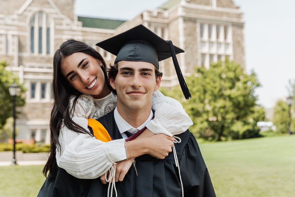 Couple celebrating graduation, woman hugging man in cap and gown outdoors on college campus.