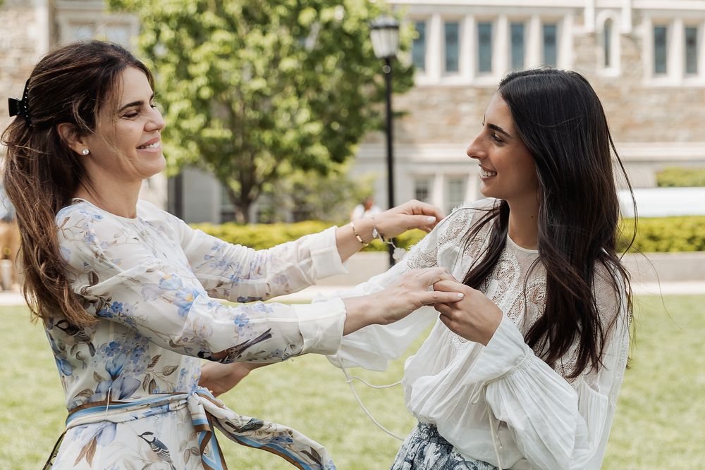 Two young women in floral dresses laugh and play together on a sunny college campus lawn.