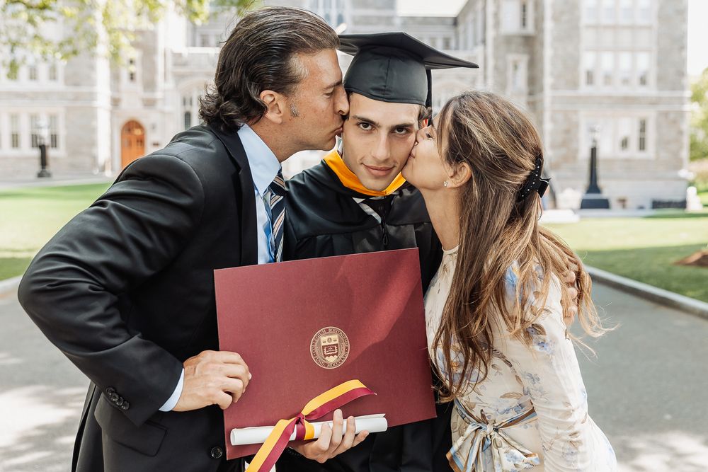 Graduate in cap and gown holds diploma while parents kiss his cheeks outdoors at university graduation ceremony.