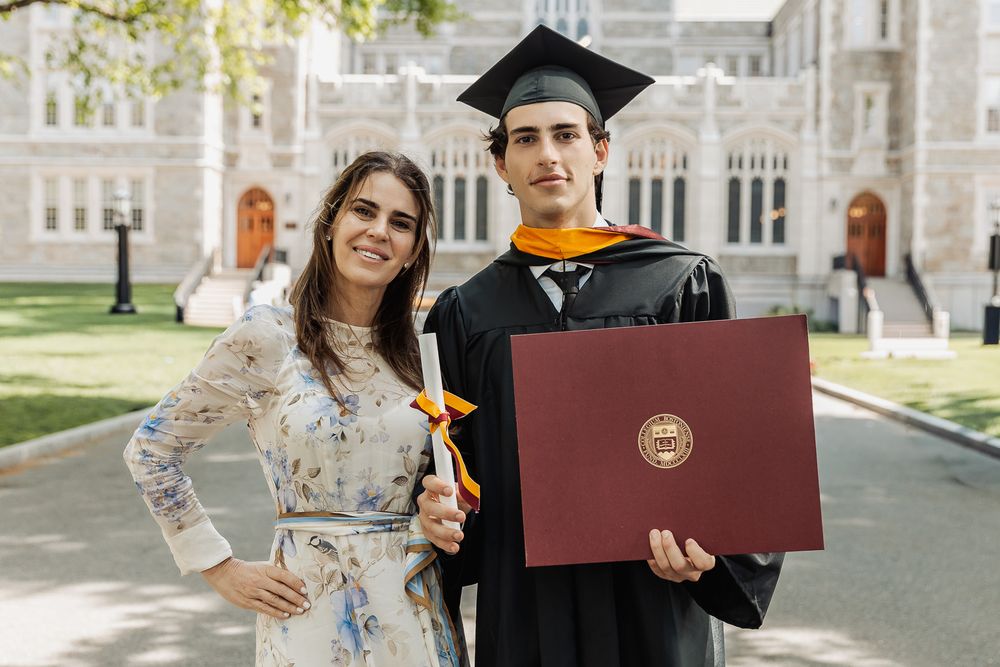 Two graduates posing outdoors with diploma in red folder, wearing cap and gown at university commencement ceremony.