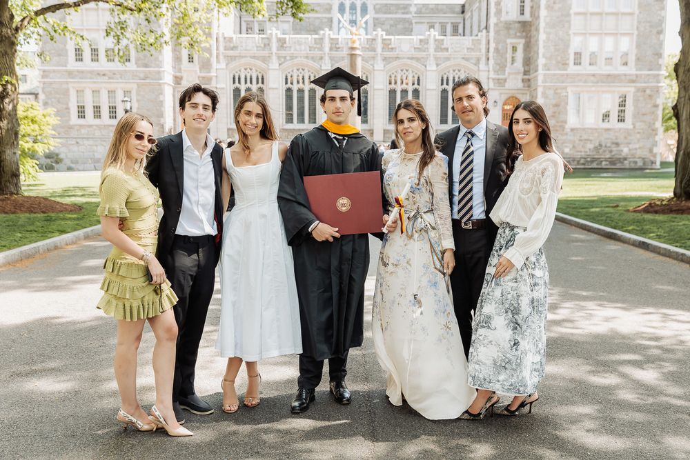 Family poses with college graduate in cap and gown holding red diploma folder outdoors at university campus.