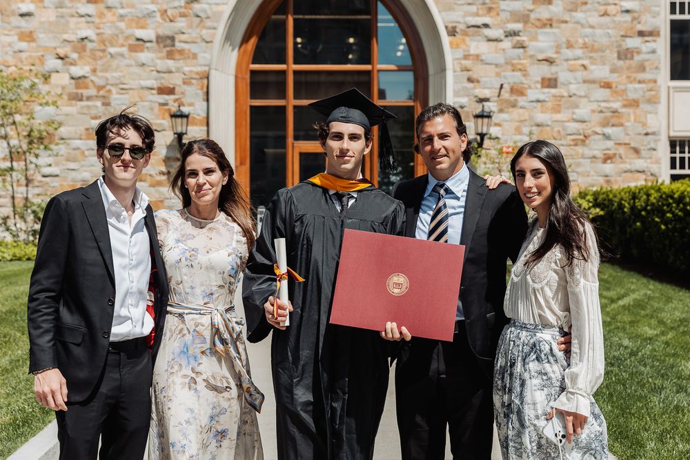 Family of five celebrates college graduation outdoors, graduate in cap and gown holds red diploma folder.