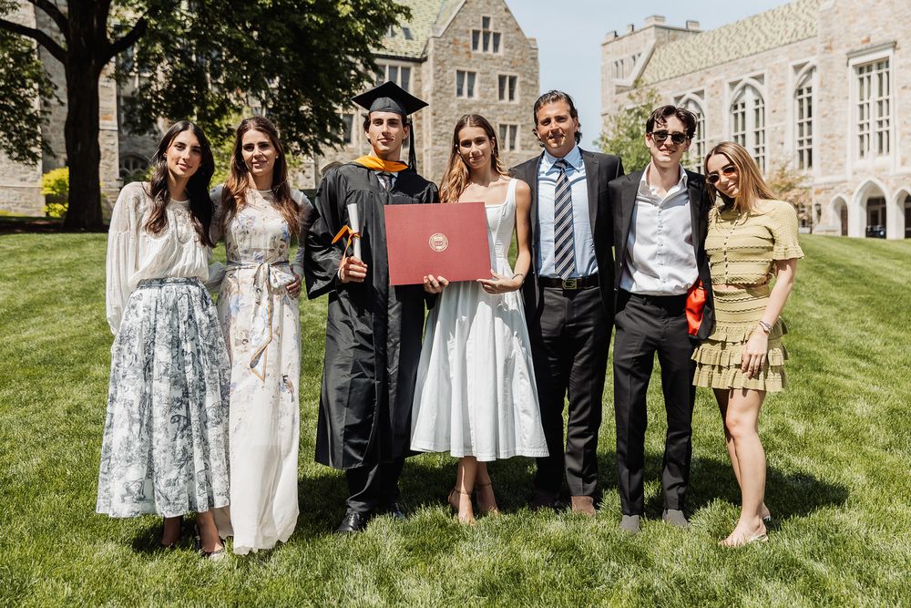 Group of seven people posing with a graduate in cap and gown holding a diploma at an outdoor university ceremony.