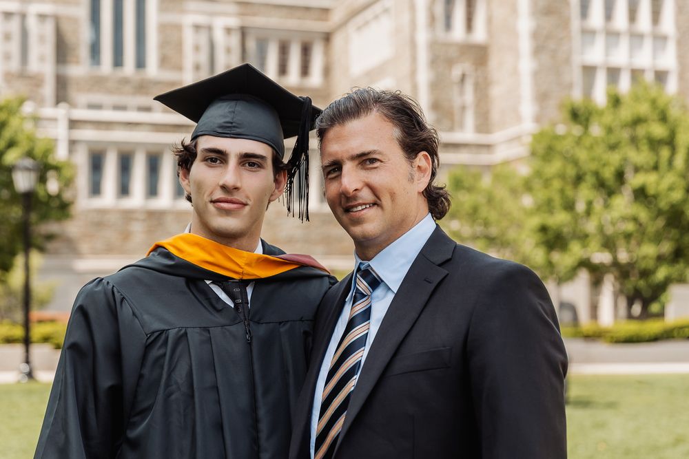 Graduate in cap and gown poses with proud father outside university building on graduation day.