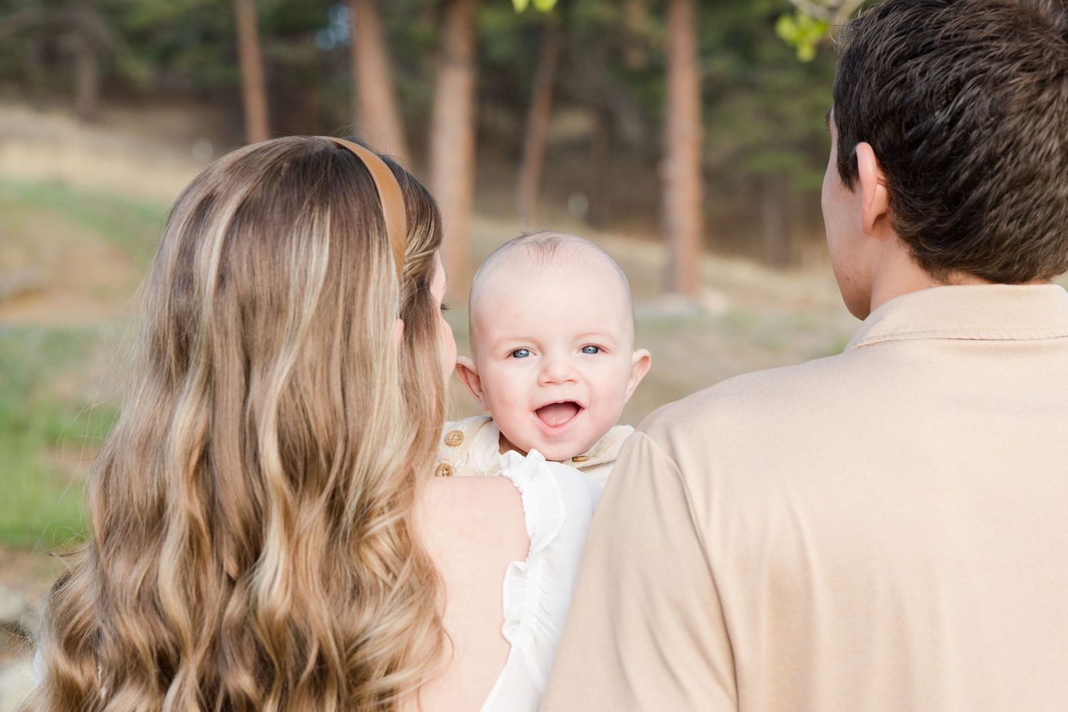 Parents holding their smiling baby outdoors among pine trees in a warm, natural family portrait.