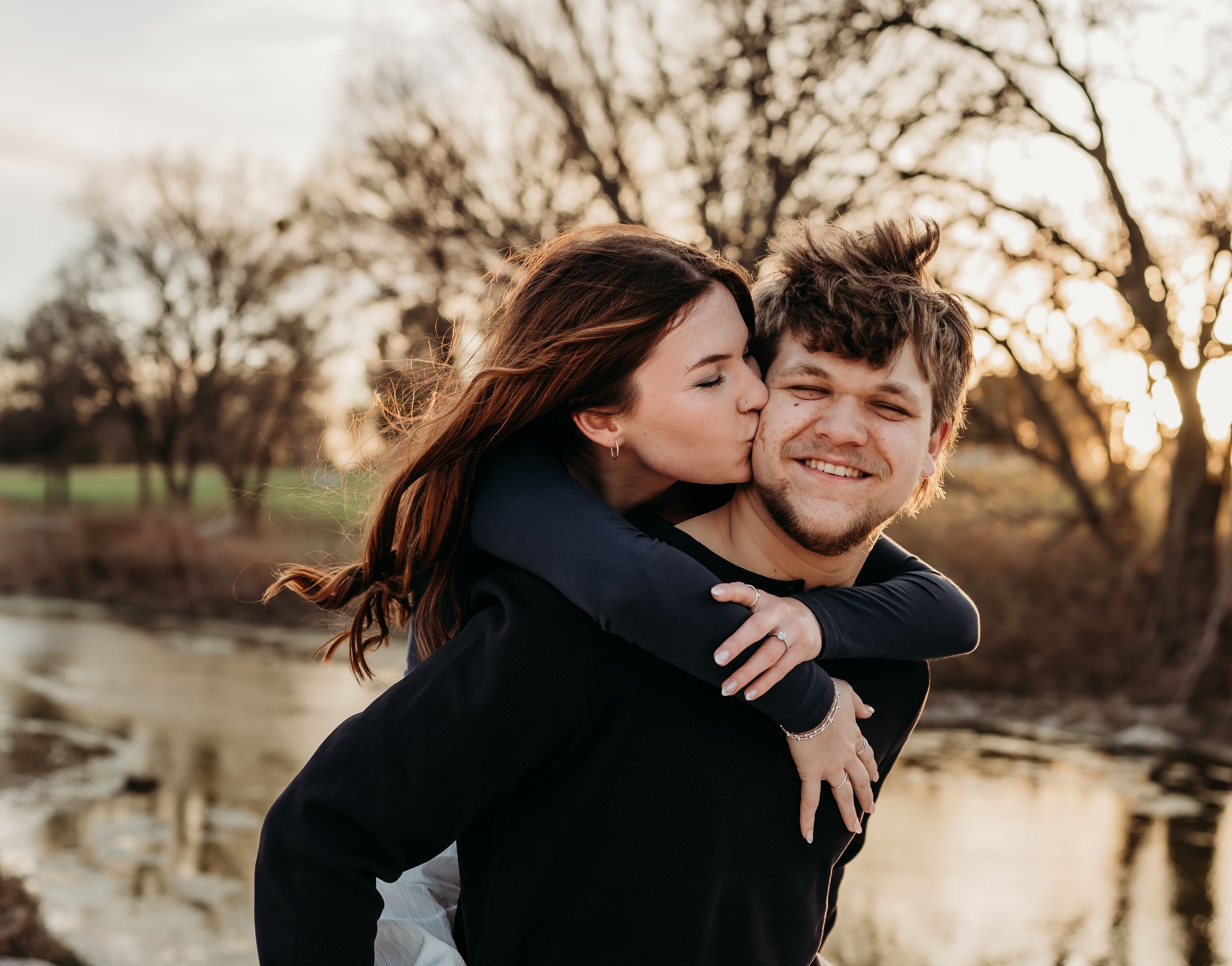 A woman kisses a smiling man on the cheek during a romantic piggyback ride outdoors near a pond at sunset.