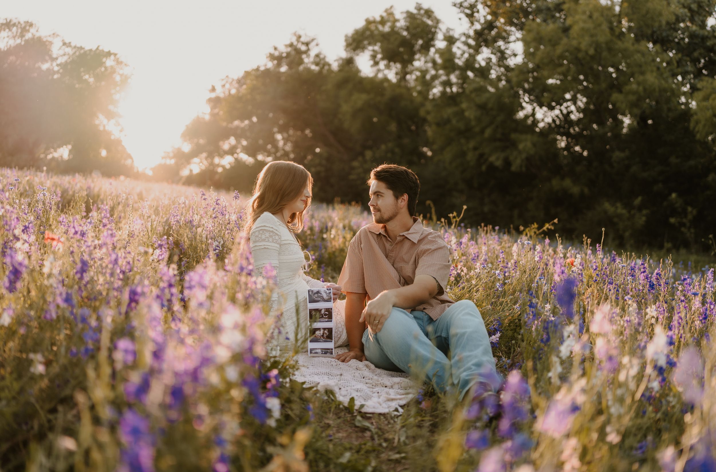 Couple sitting in a sunlit purple wildflower field during golden hour, sharing an intimate moment outdoors.