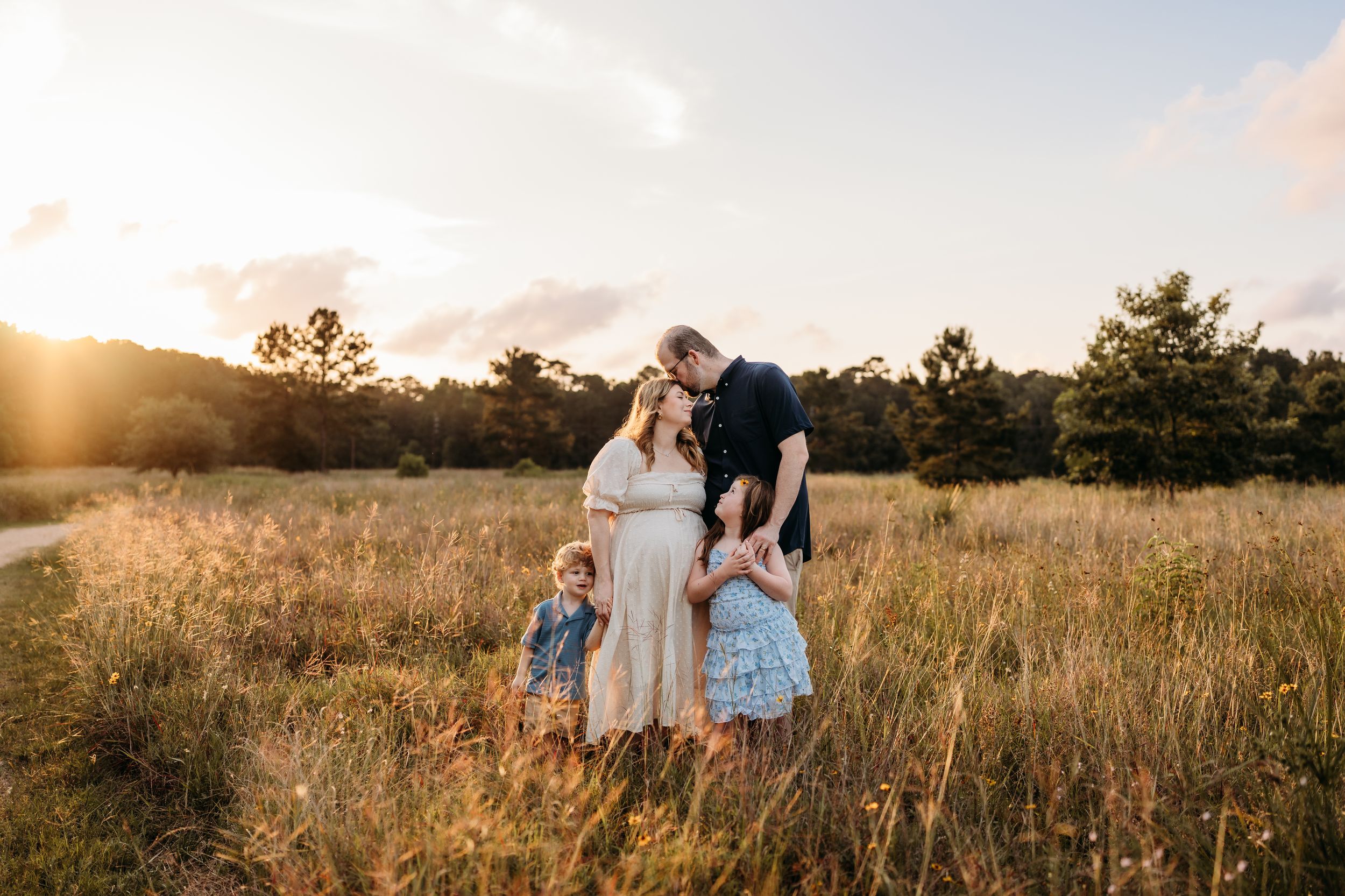 Family of four poses in golden field at sunset, pregnant mother and father with two young children.