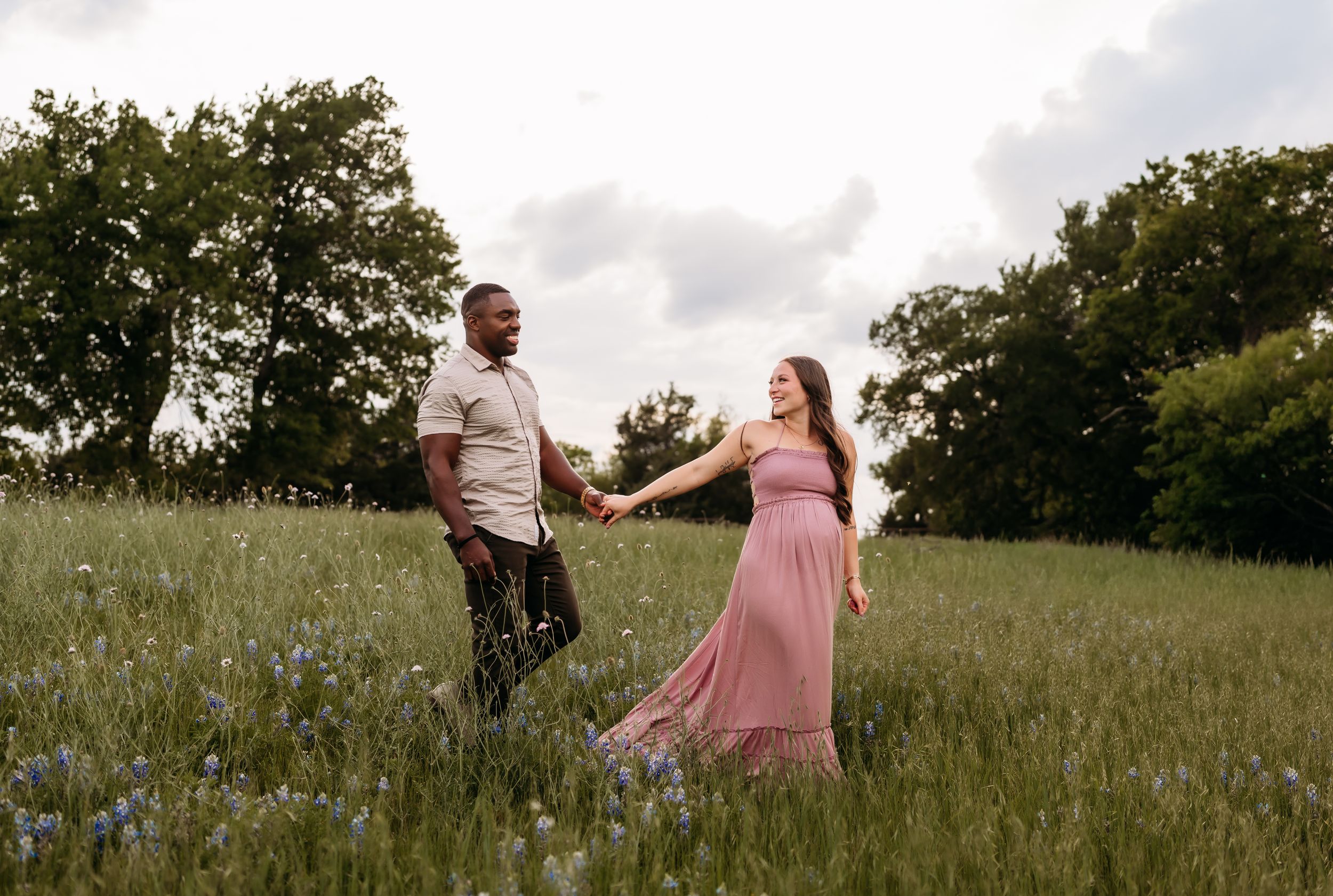 Couple holding hands walking through a wildflower meadow, woman in pink maxi dress, man in casual attire.