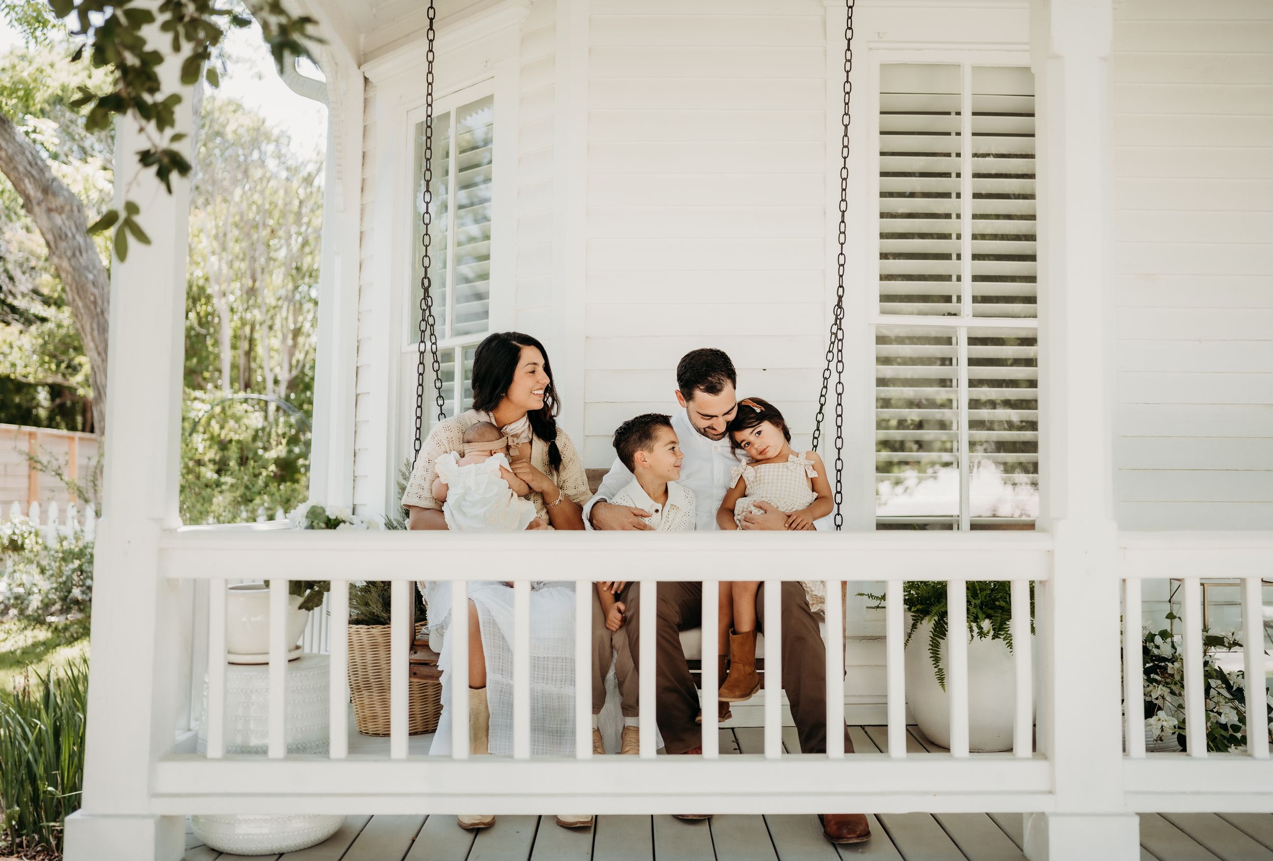 Family of four sitting on a white porch swing, smiling together outside a charming white home.