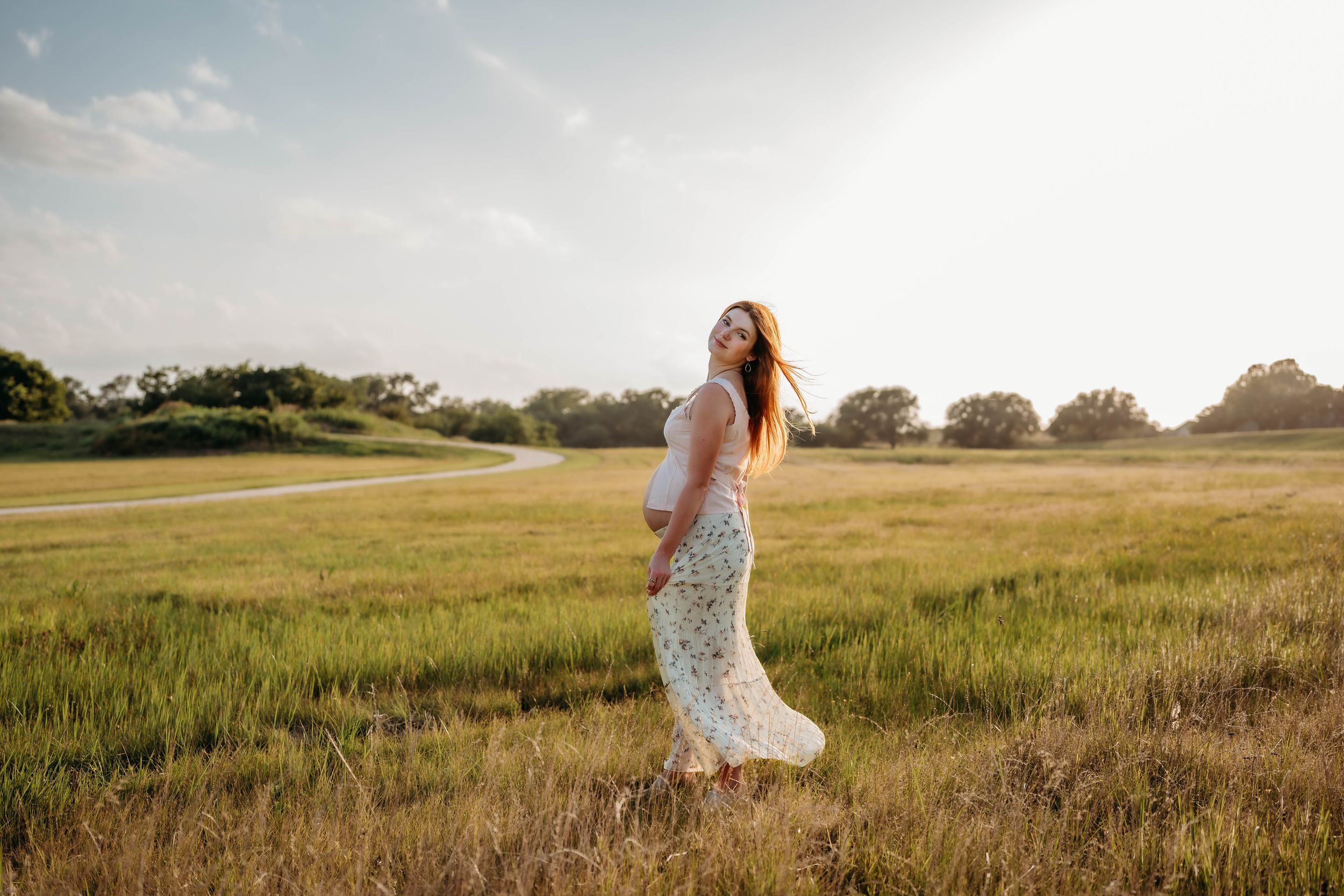 Pregnant woman in floral dress standing in golden sunlit field, cradling baby bump during outdoor maternity photoshoot.