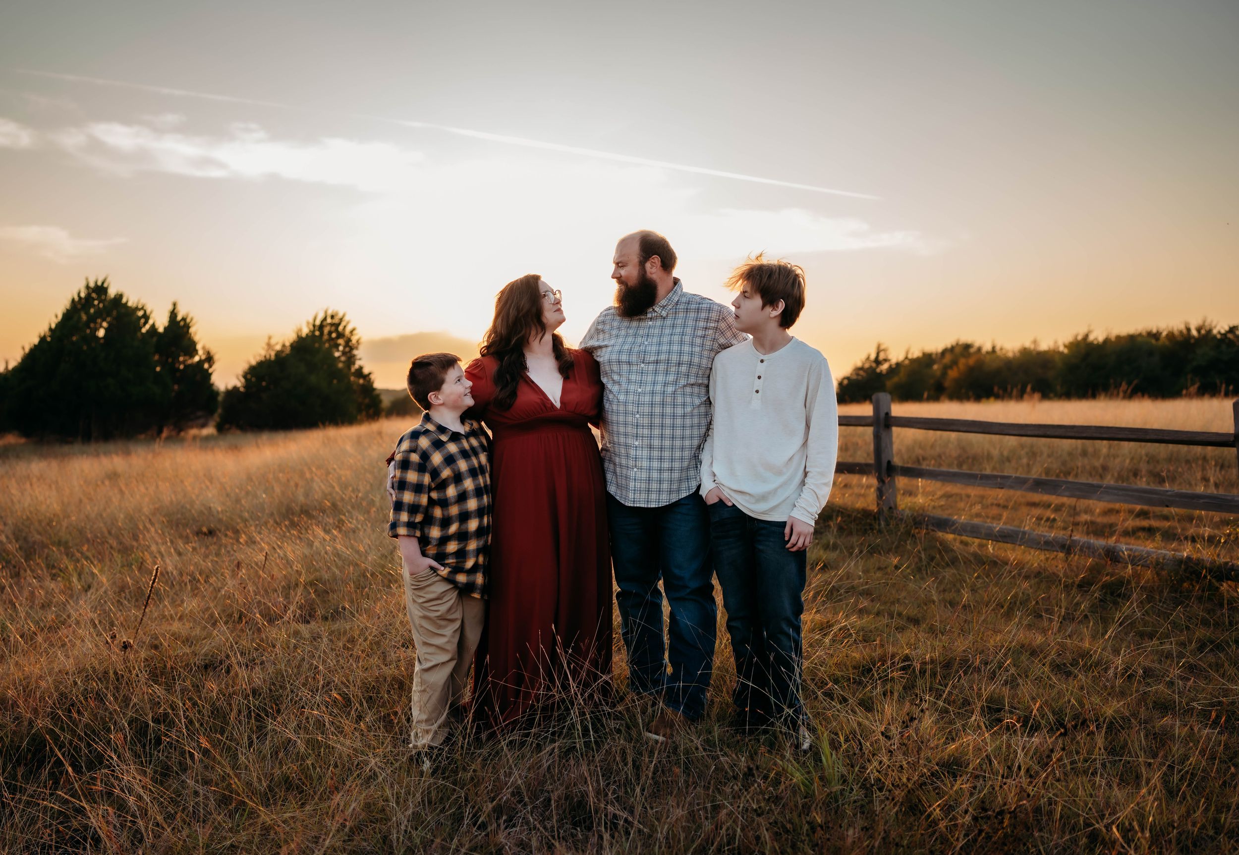 Family of four posing in golden field at sunset, mother in red dress, father and two sons smiling outdoors.
