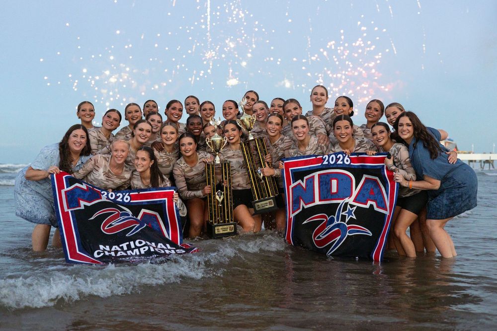 NDA National Champions 2026 dance team celebrates with trophy and banners while standing in ocean waves.