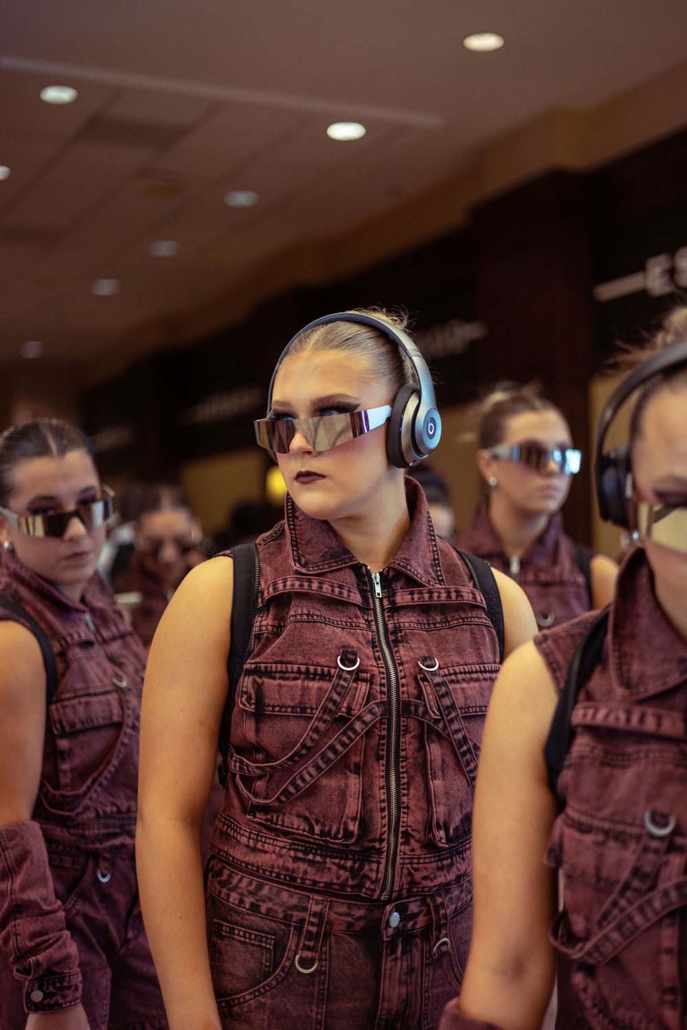 Group in matching maroon denim vests and sunglasses stand listening with headphones during a performance.