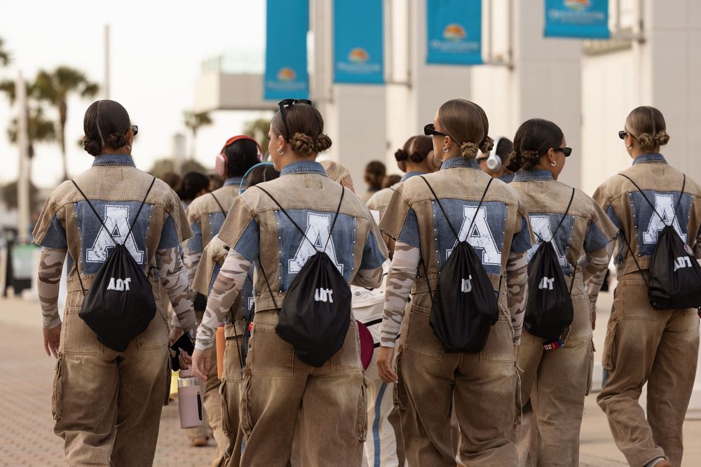 Group of women in matching tan uniforms with black drawstring backpacks walking outdoors near a building.