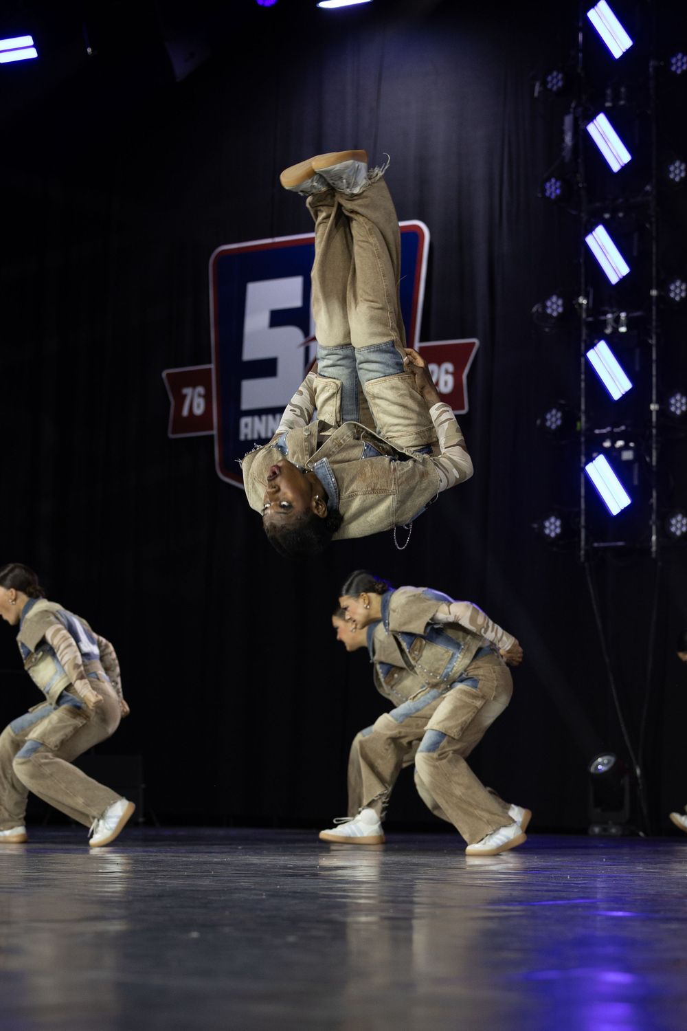 Acrobatic dancer performing an inverted flip while teammates crouch on stage at a 50th anniversary dance competition.
