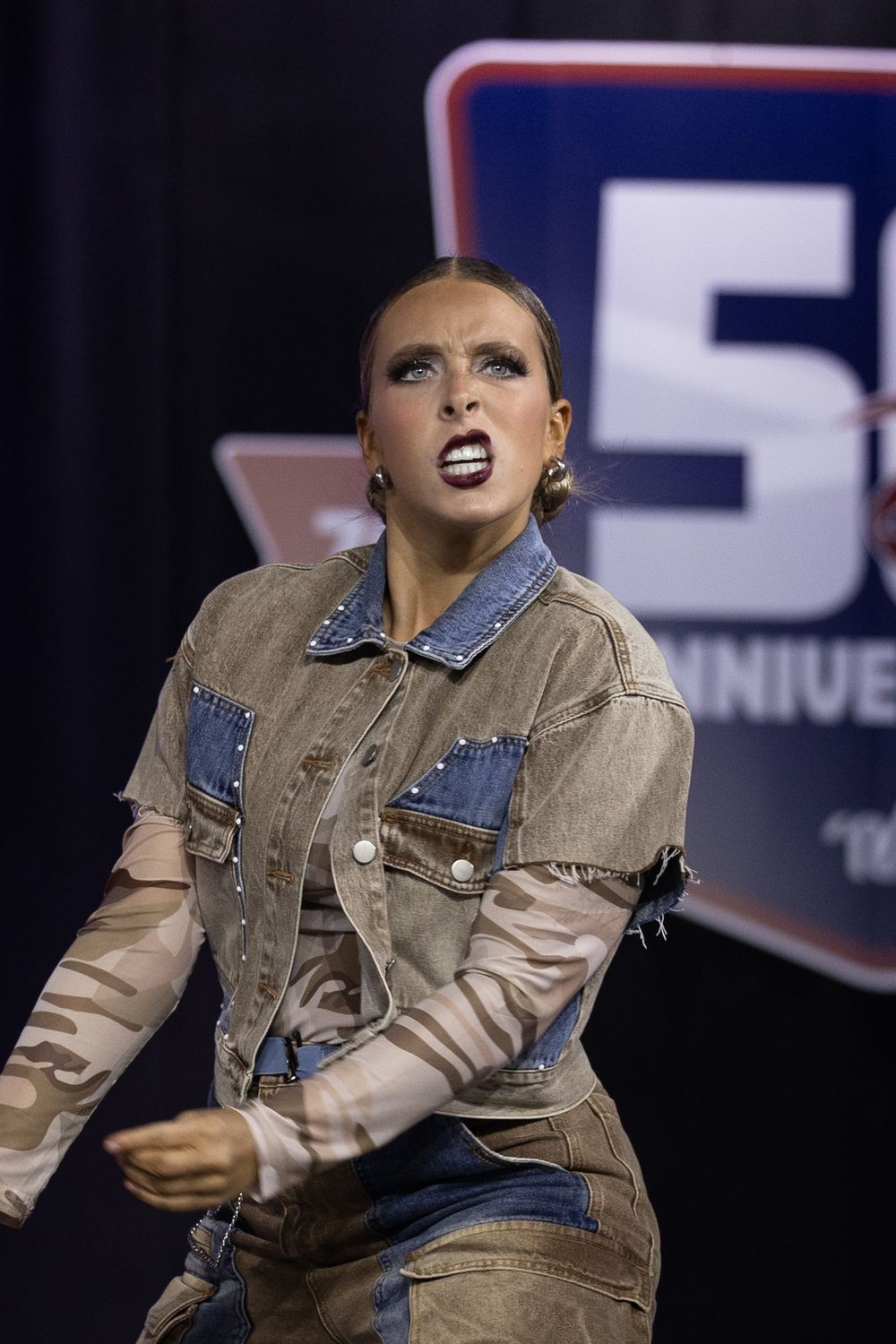Female dancer in denim jacket with camo sleeves performs on stage at a 50th anniversary competition event.