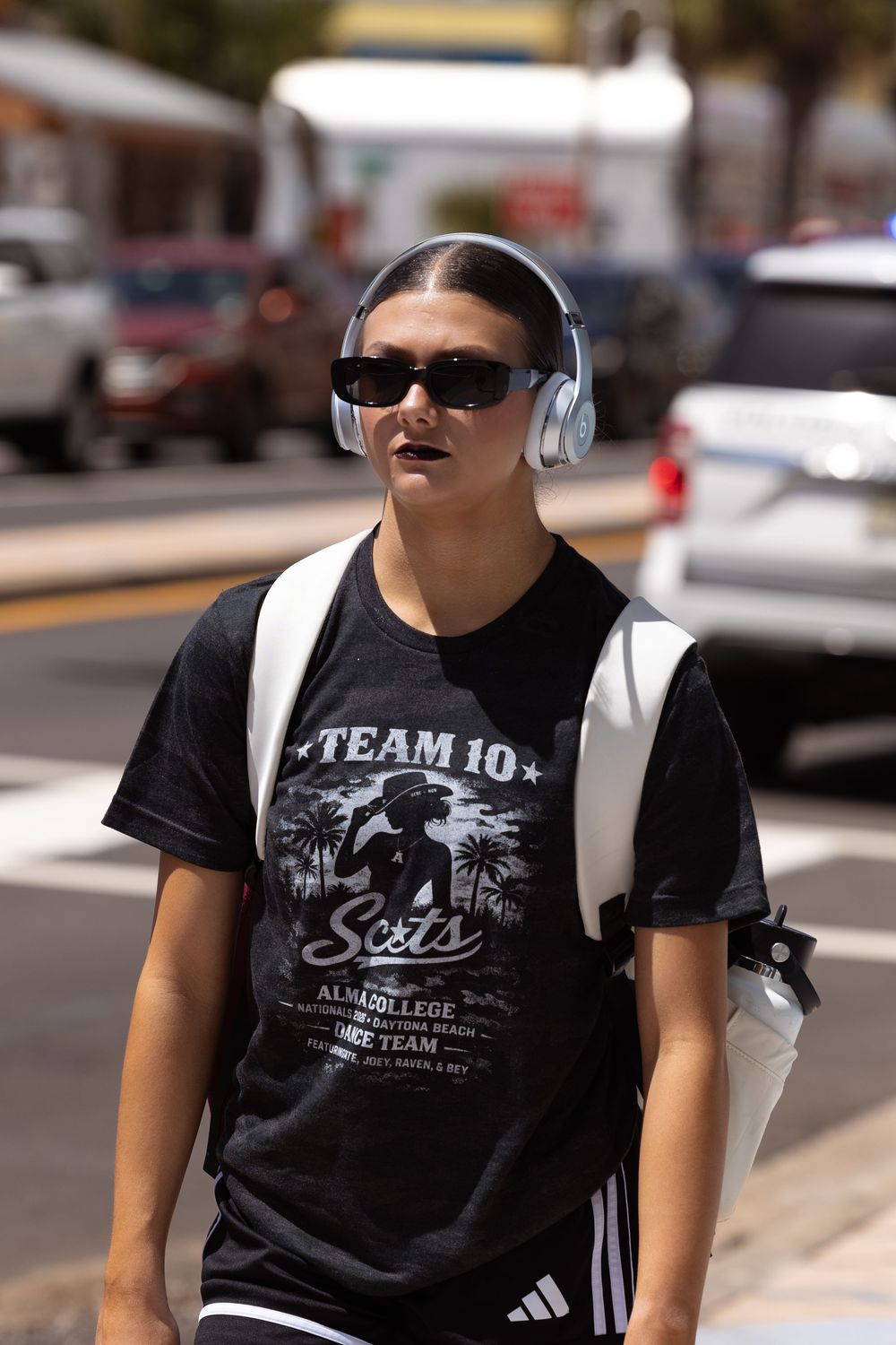 Young person wearing Team 10 black tee, white headphones, sunglasses, and Adidas backpack walking outdoors.