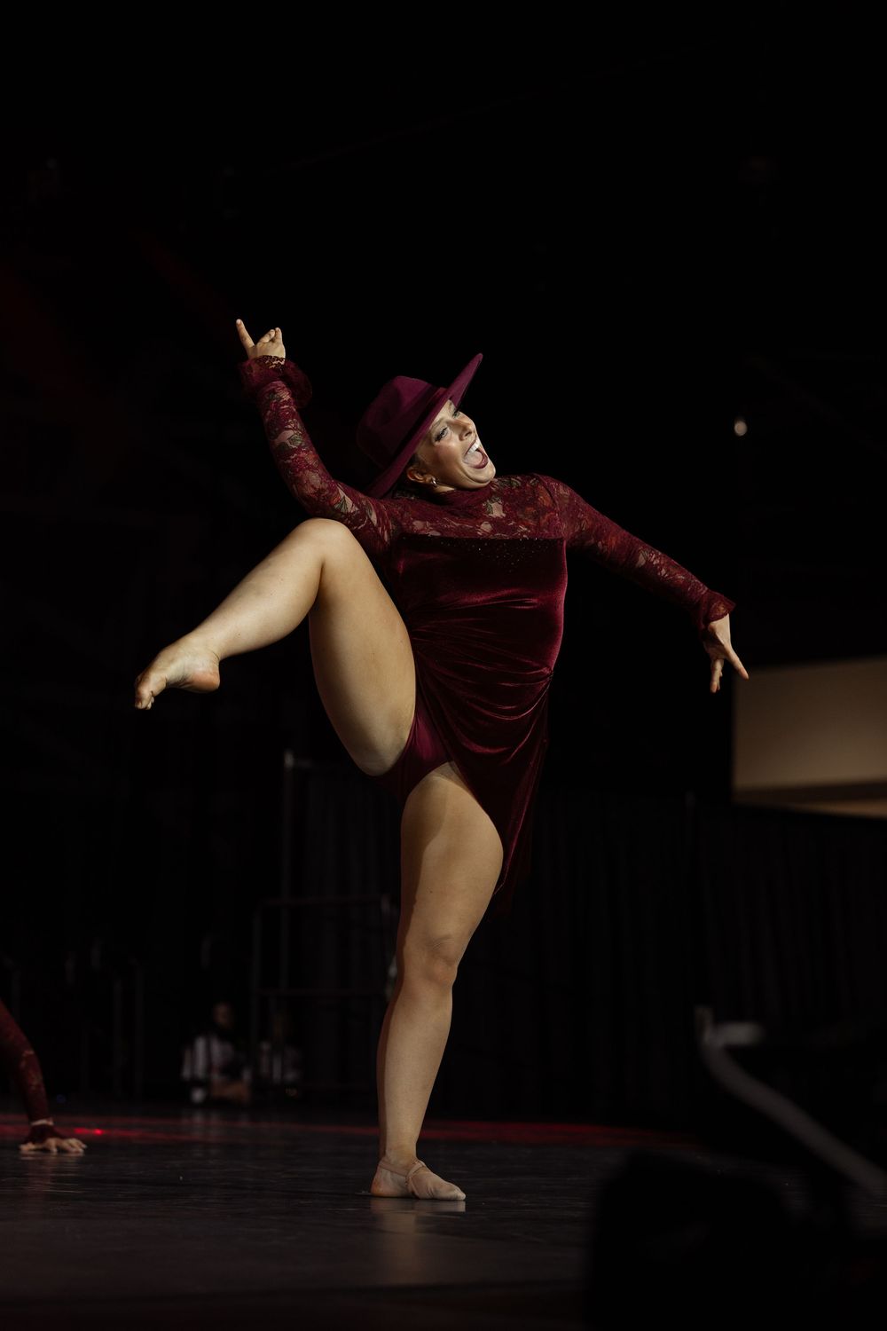 A dancer in a burgundy velvet leotard and hat performs a high kick on stage under dramatic lighting.