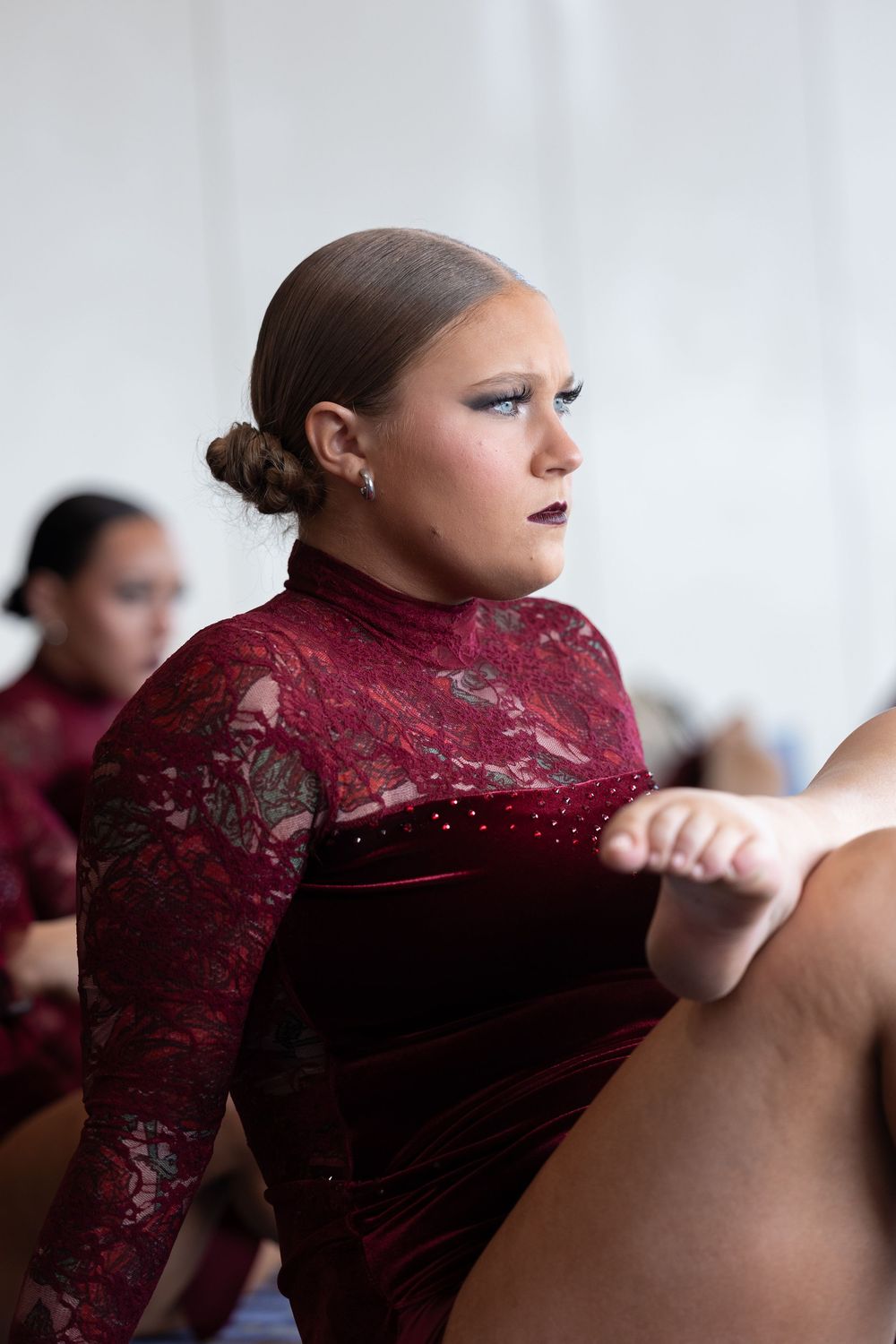 Young female dancer in a burgundy lace leotard stretching during a dance competition or rehearsal.