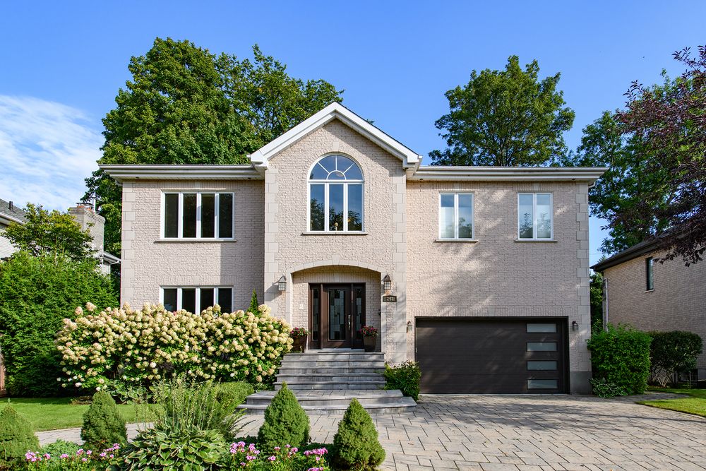 Elegant two-story brick home with arched window, dark garage door, manicured shrubs, and paver driveway surrounded by lush trees.
