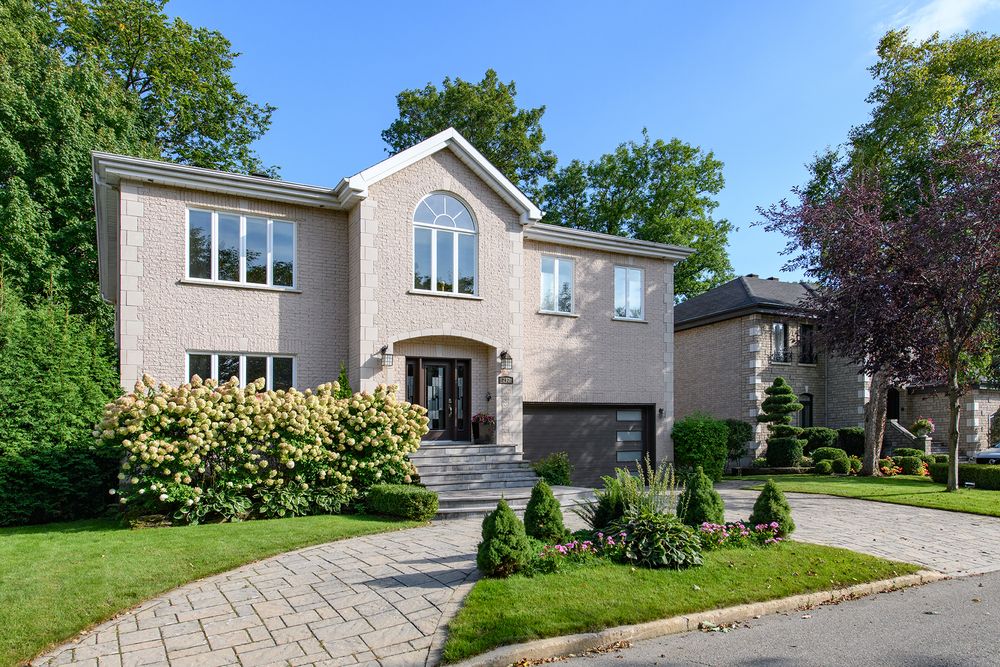 Elegant two-story brick home with arched window, manicured landscaping, and paver driveway surrounded by lush green trees.
